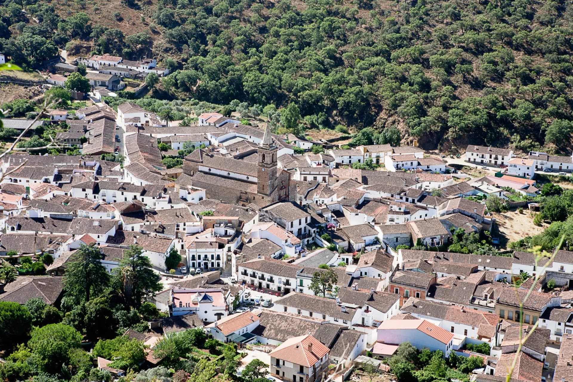 Aerial view of white village with terracotta roofs nestled against a steep, tree-covered hillside in Alájar, Huelva.