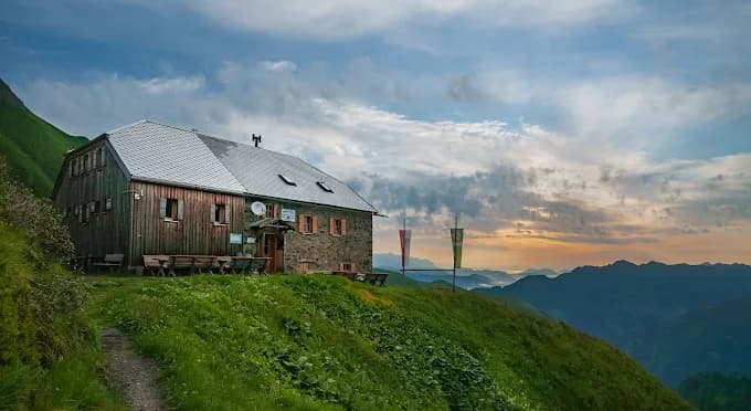 Gleiwitzer Hütte mountain refuge on grassy slope overlooking layered mountain ranges at sunset.