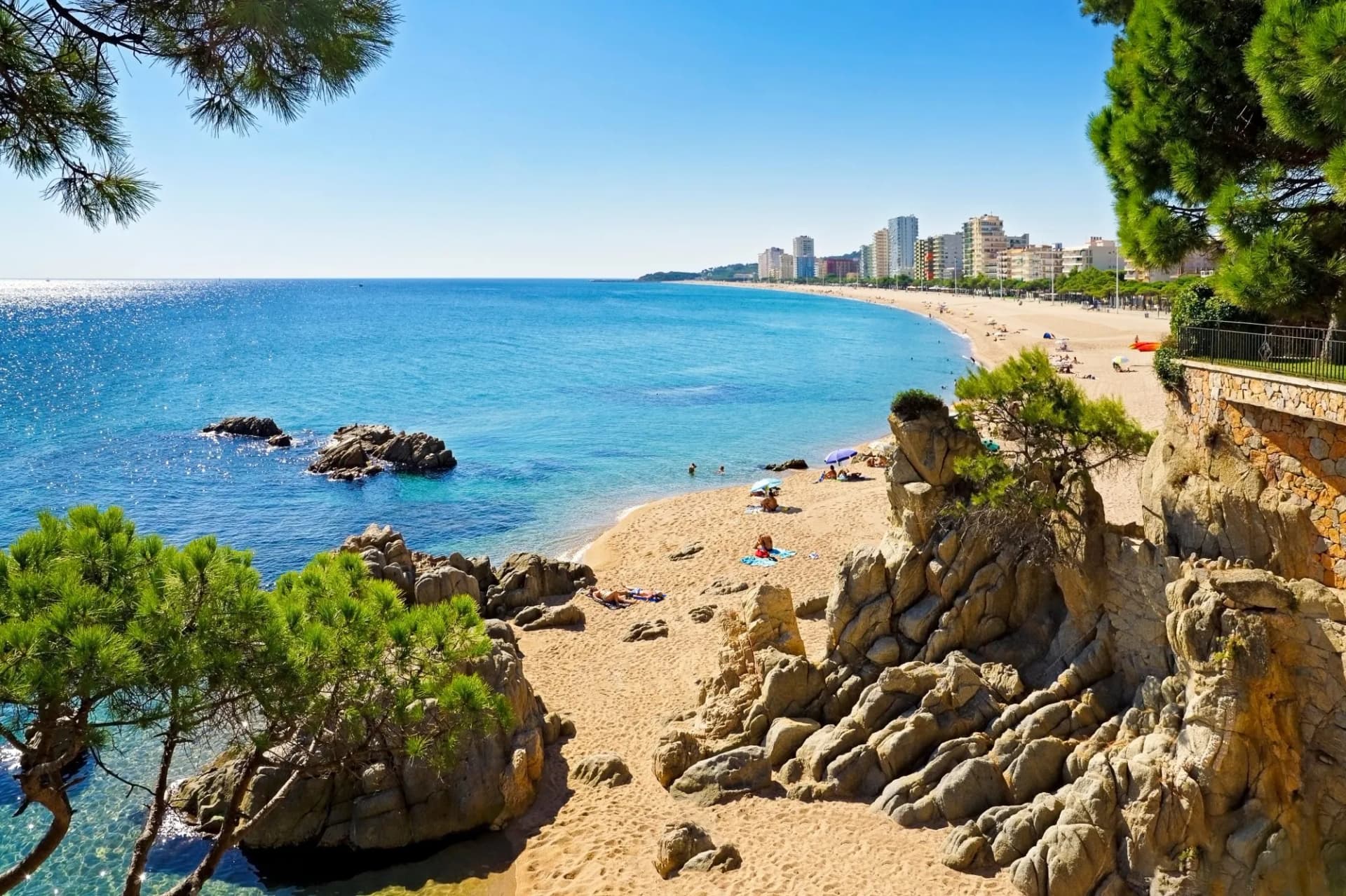 Beach in Platja d'Aro, Spain, with turquoise sea, rocky foreground, and city skyline.