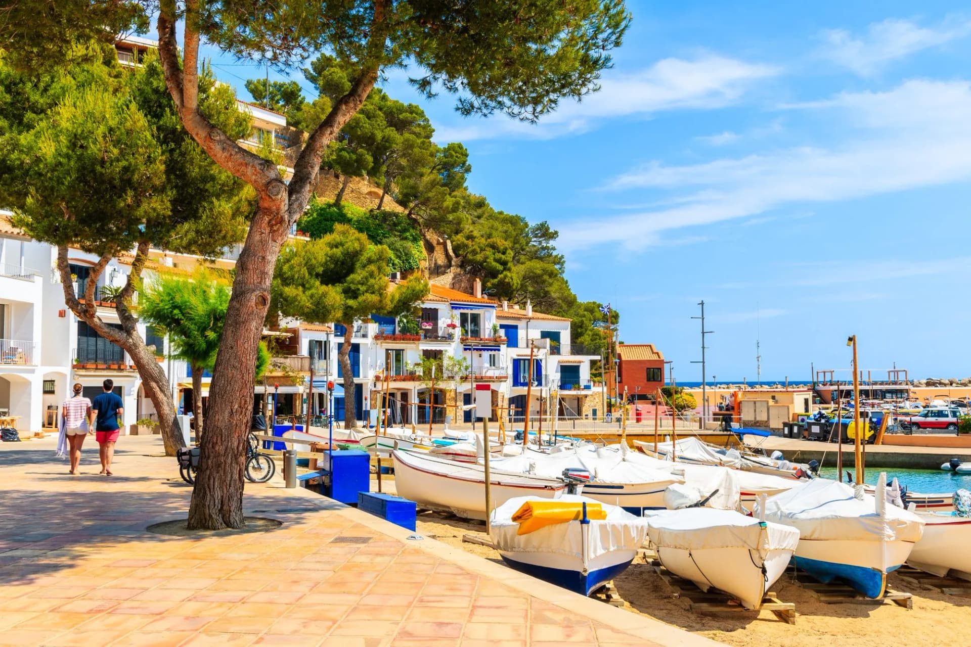Couple walking past covered fishing boats on beach in Llafranc village, Costa Brava, Spain.