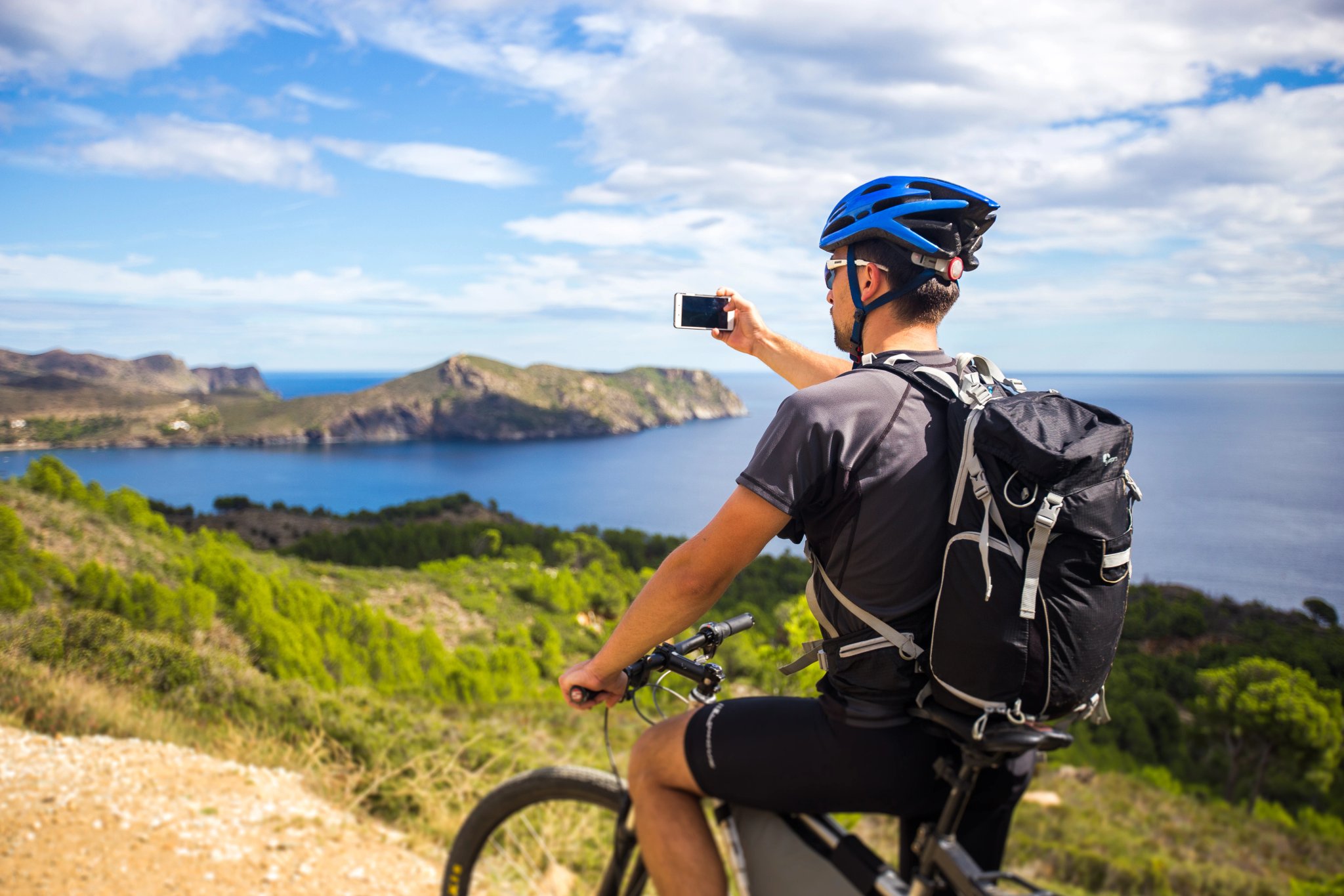 junger Mann auf Mountainbike-Strecken in Spanien und macht ein Foto mit weißem Handy im Hintergrund des Mittelmeers an der zerklüfteten Küste der Costa Brava. Mit blauem Helm auf dem Kopf