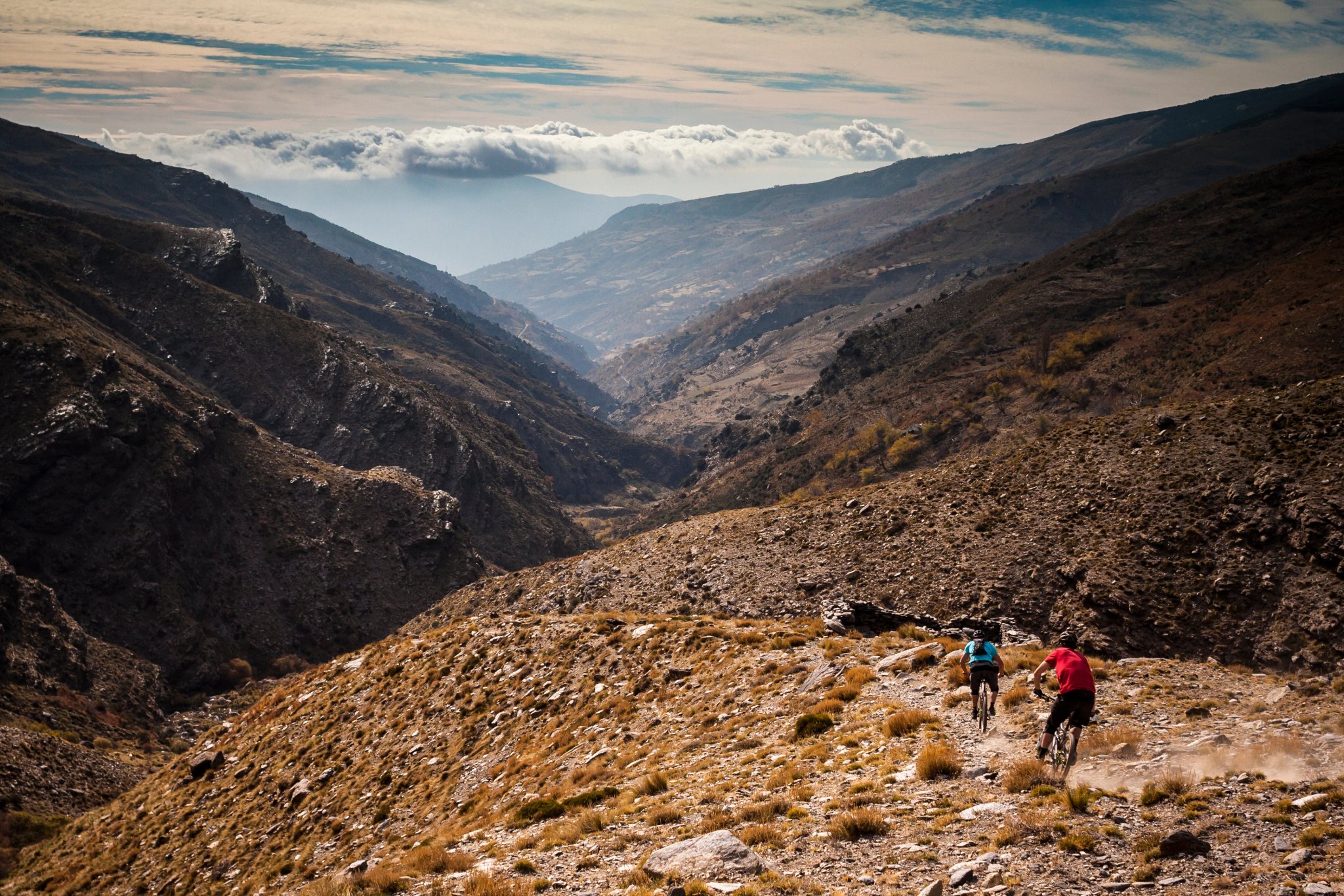SIERRA NEVADA, SPANIEN. Två mountainbikers cyklar nerför en grov singletrack-stig i en karg avlägsen dal med dramatisk himmel och dimma.