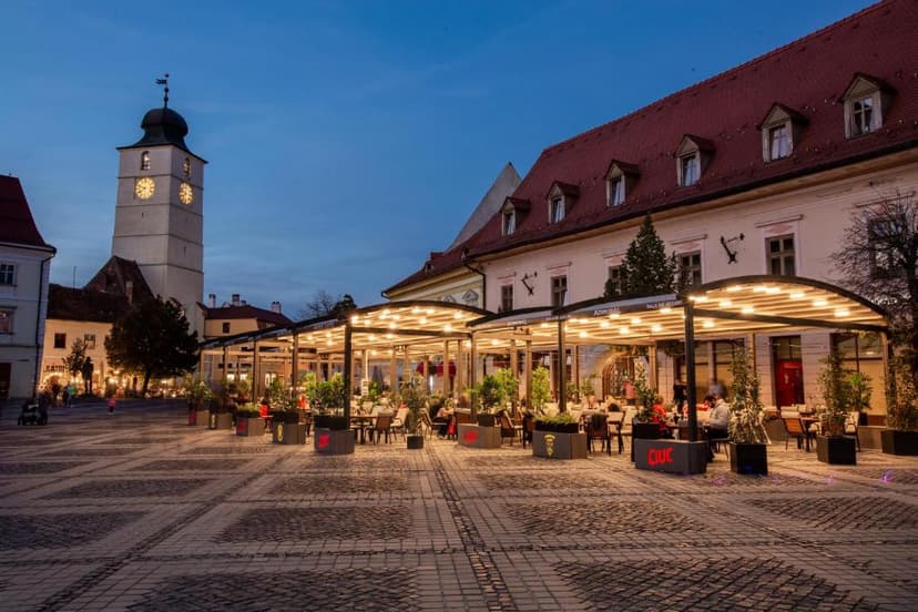 Outdoor cafe seating under illuminated pergolas in a historic square with a clock tower.