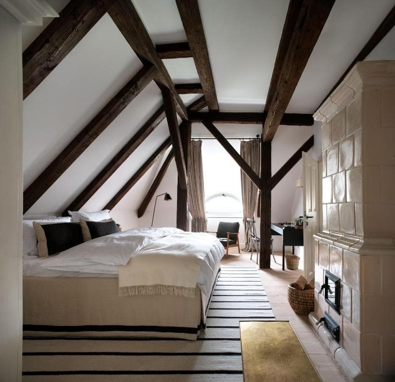 Attic bedroom with exposed dark wood beams, white bedding, and a tiled stove in Bethlen Estates.