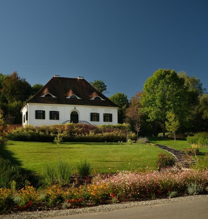 White historic estate house with dark roof surrounded by lush green lawn and colorful flower beds.
