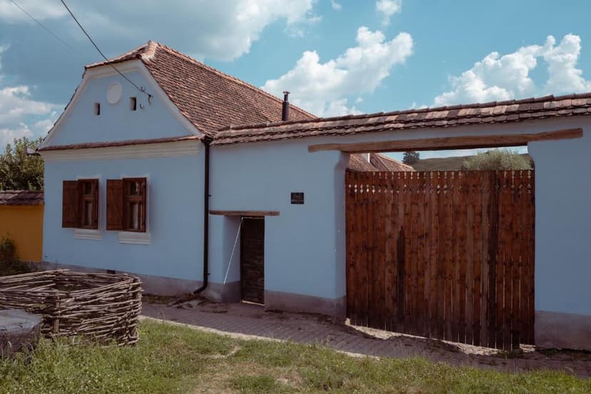 Light blue house with terracotta roof and large wooden gate under a cloudy blue sky.