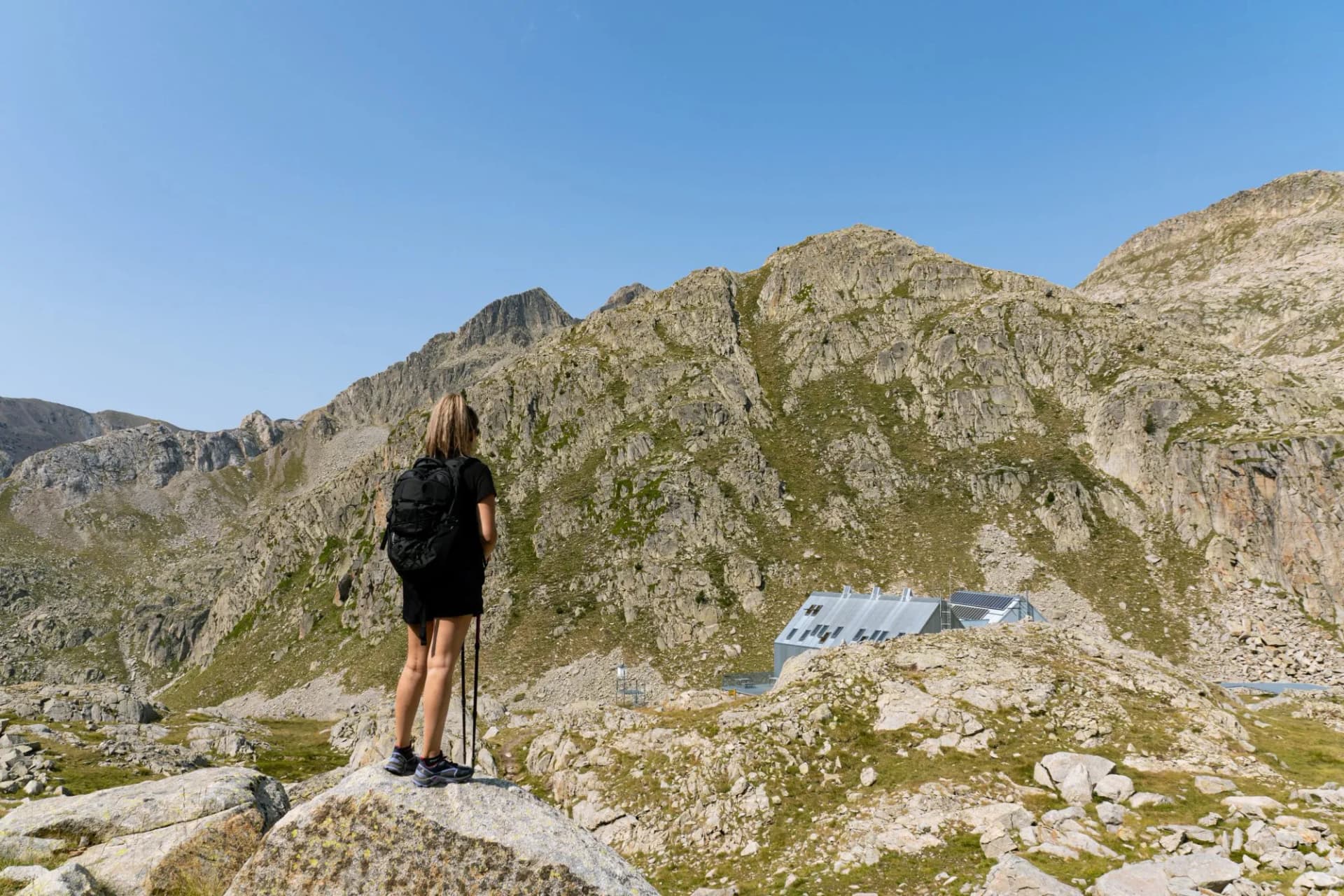 Hiker with backpack and poles overlooking rocky mountains and a modern refuge building.