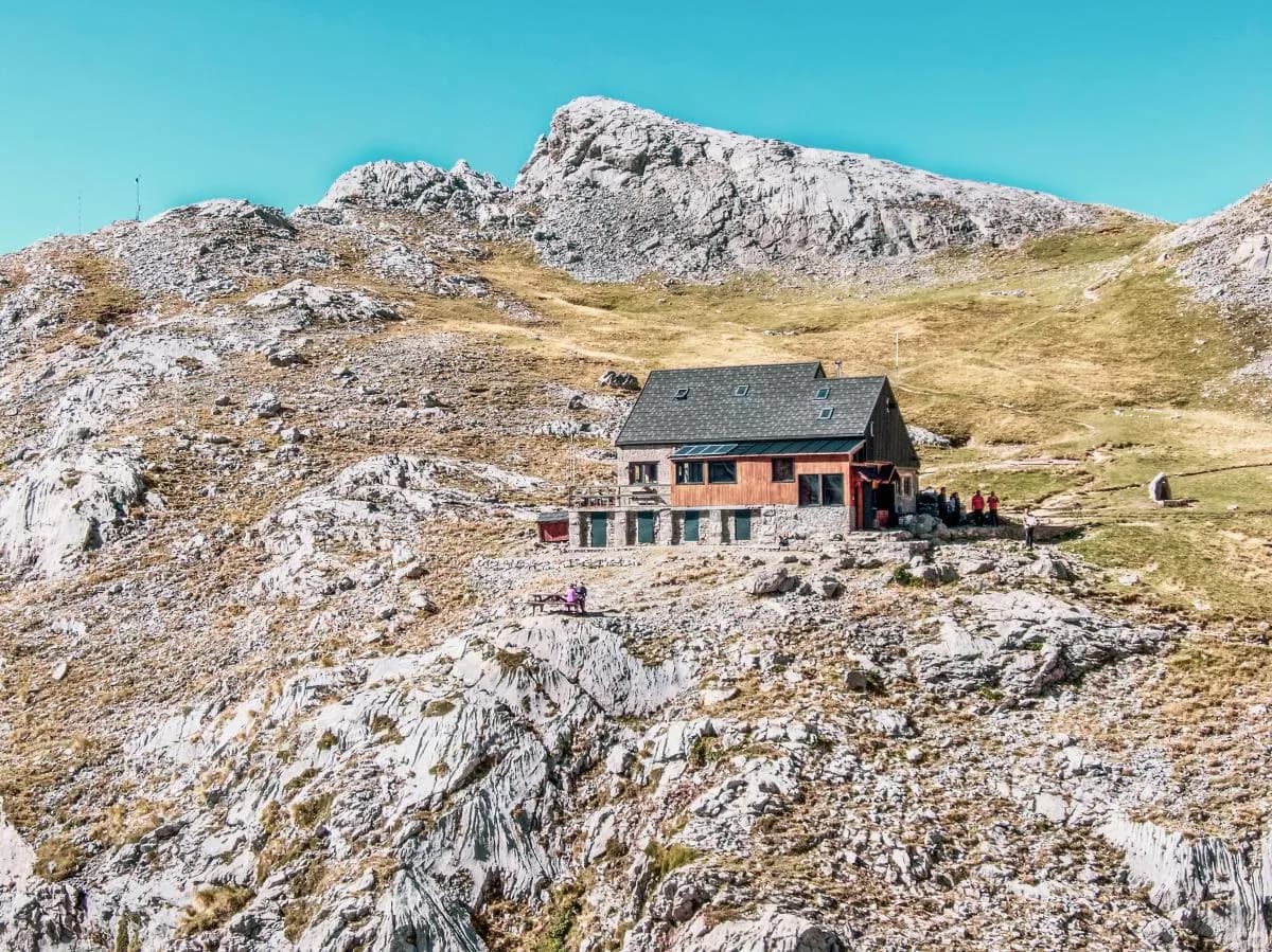 Mountain refuge Refugi Collado Jermoso on rocky terrain beneath a large peak under a clear blue sky.