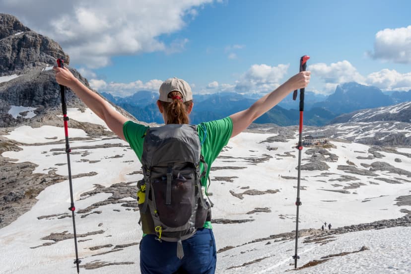 Woman with a backpack raises her hands to the sky after successfully hiking to the top of snow capped mountains. Altopiano delle Pale di San Martino, San Martino di Castrozza - Trentino, Italy