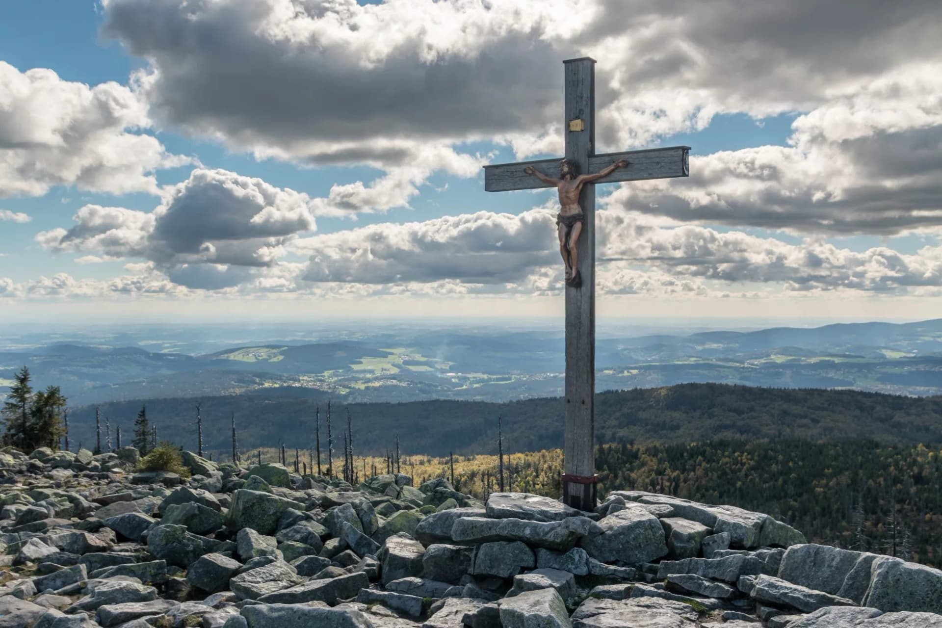 Summit cross on rocky peak overlooking forested hills under dramatic cloudy sky, Lusen-Hill