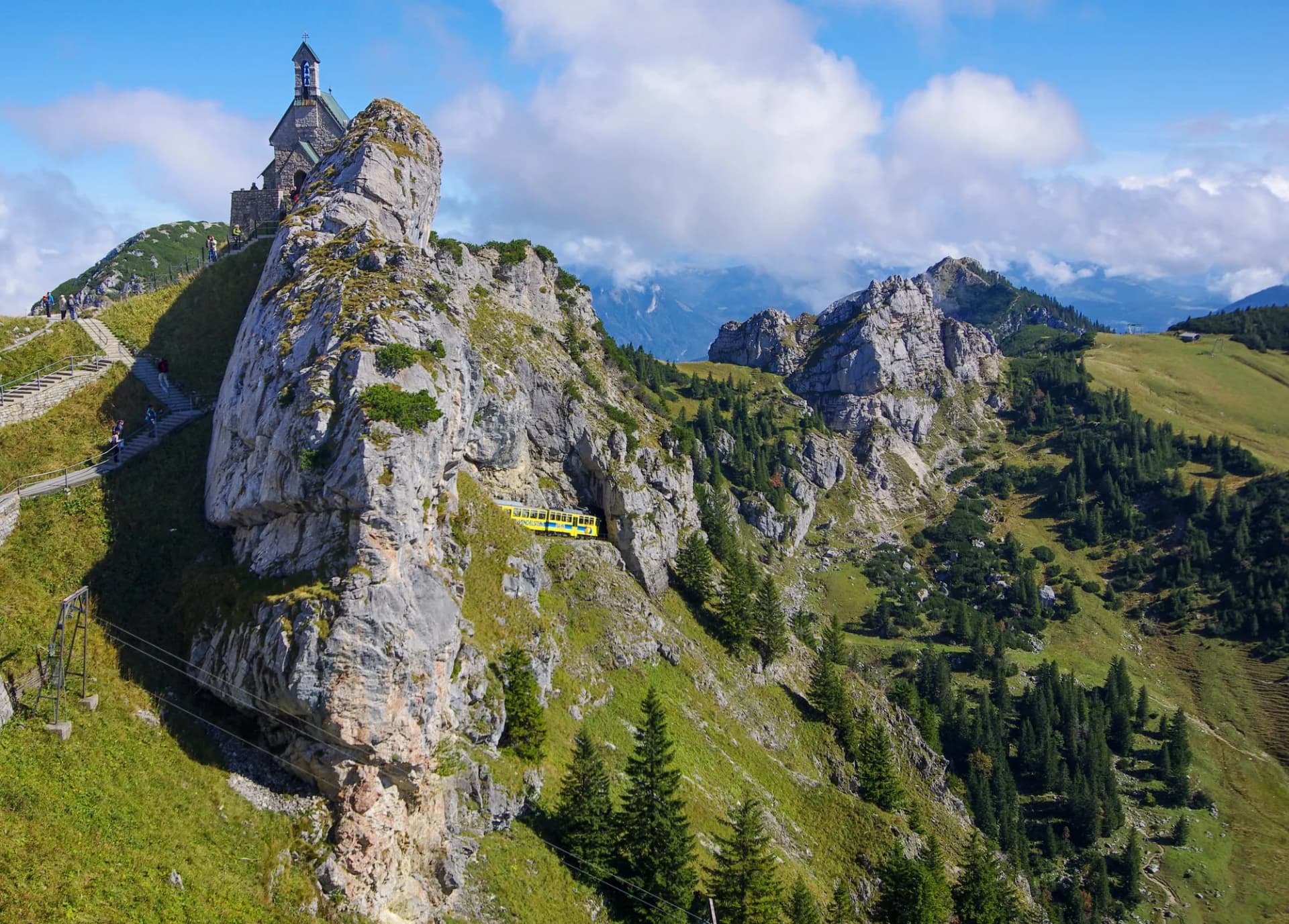 Wendelstein cogwheel train on mountain slope near chapel under blue sky with clouds.