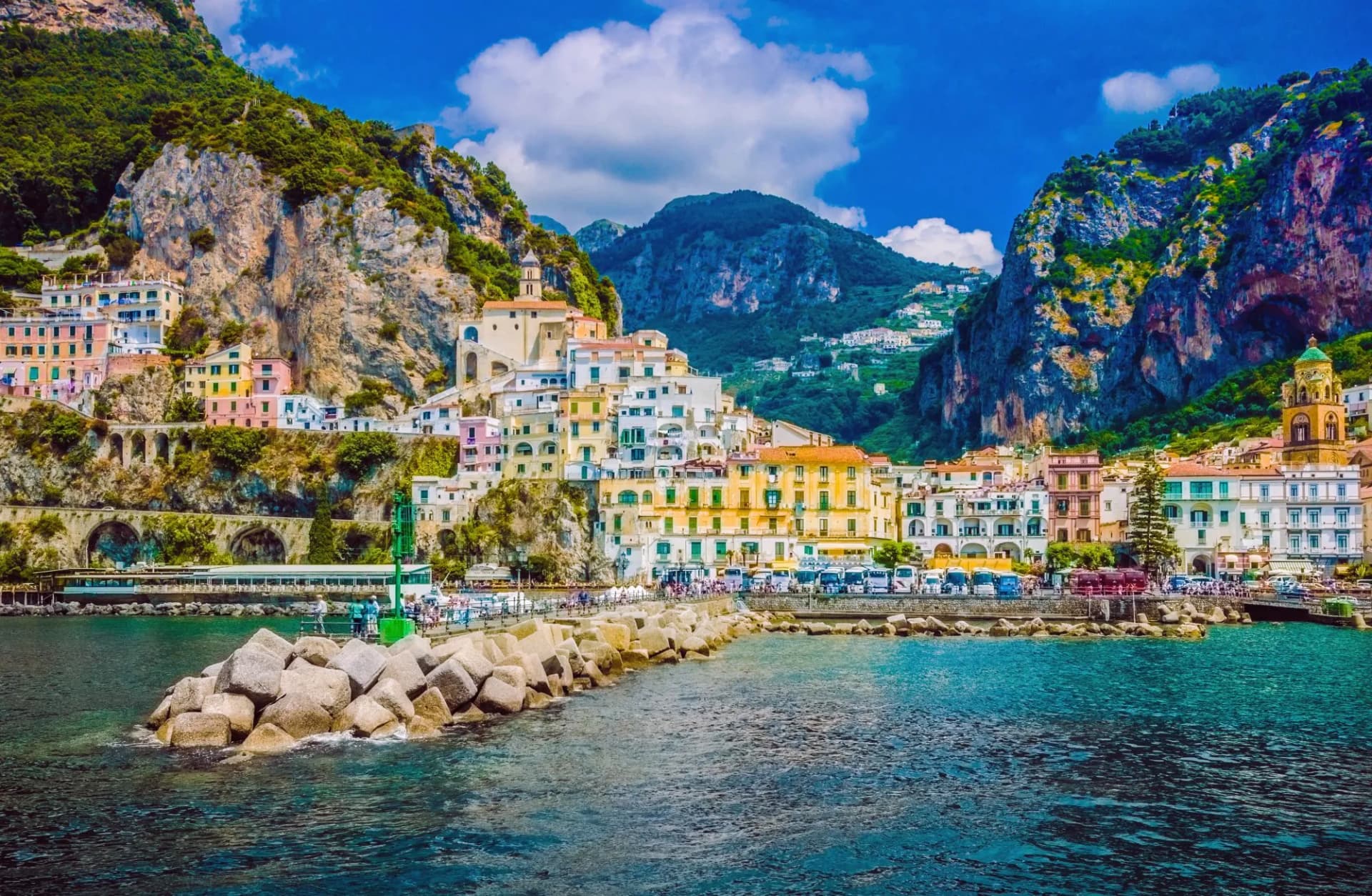Amalfi village with colorful houses on steep slopes above turquoise sea and rocky breakwater.
