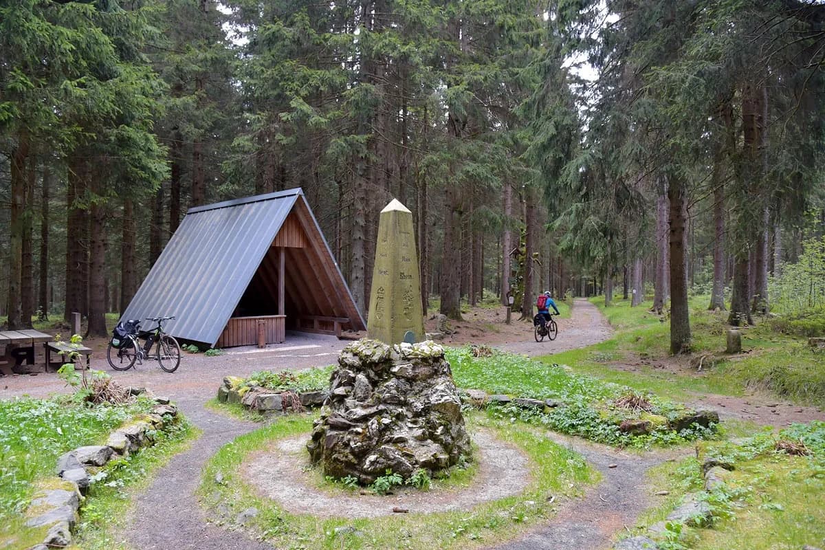 Bicyclist riding past a stone monument and shelter in a dense pine forest.