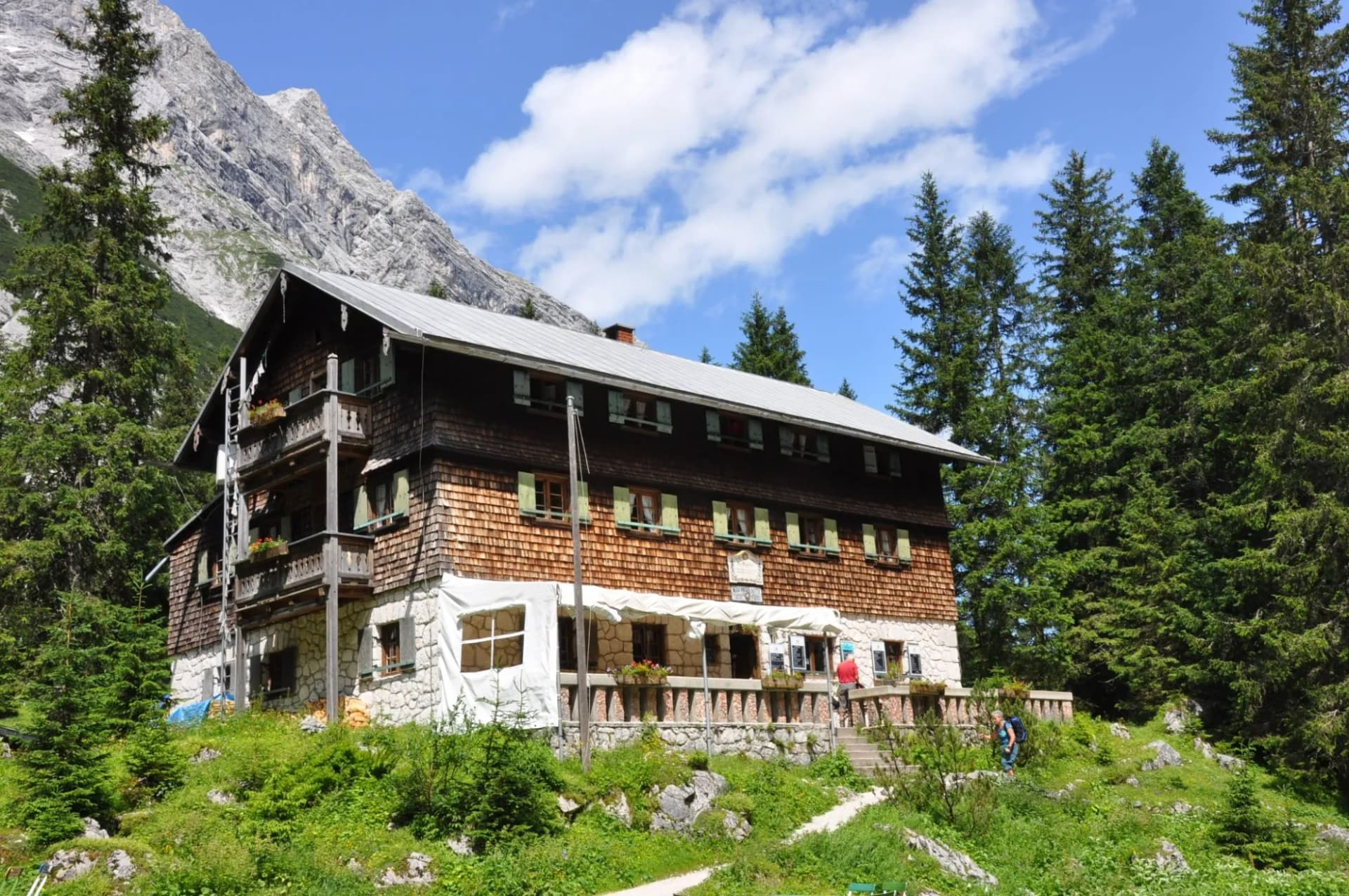 Reintalanger hut with wooden facade and stone base nestled in green mountainside under blue sky.