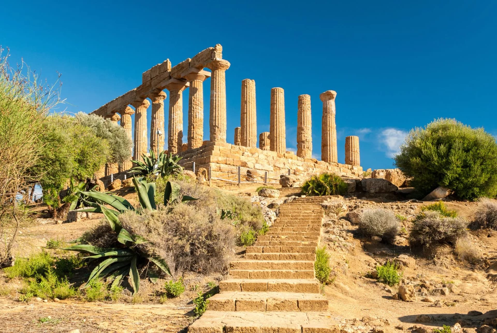 Temple of Juno ruins atop hill with stone steps and arid vegetation under blue sky.