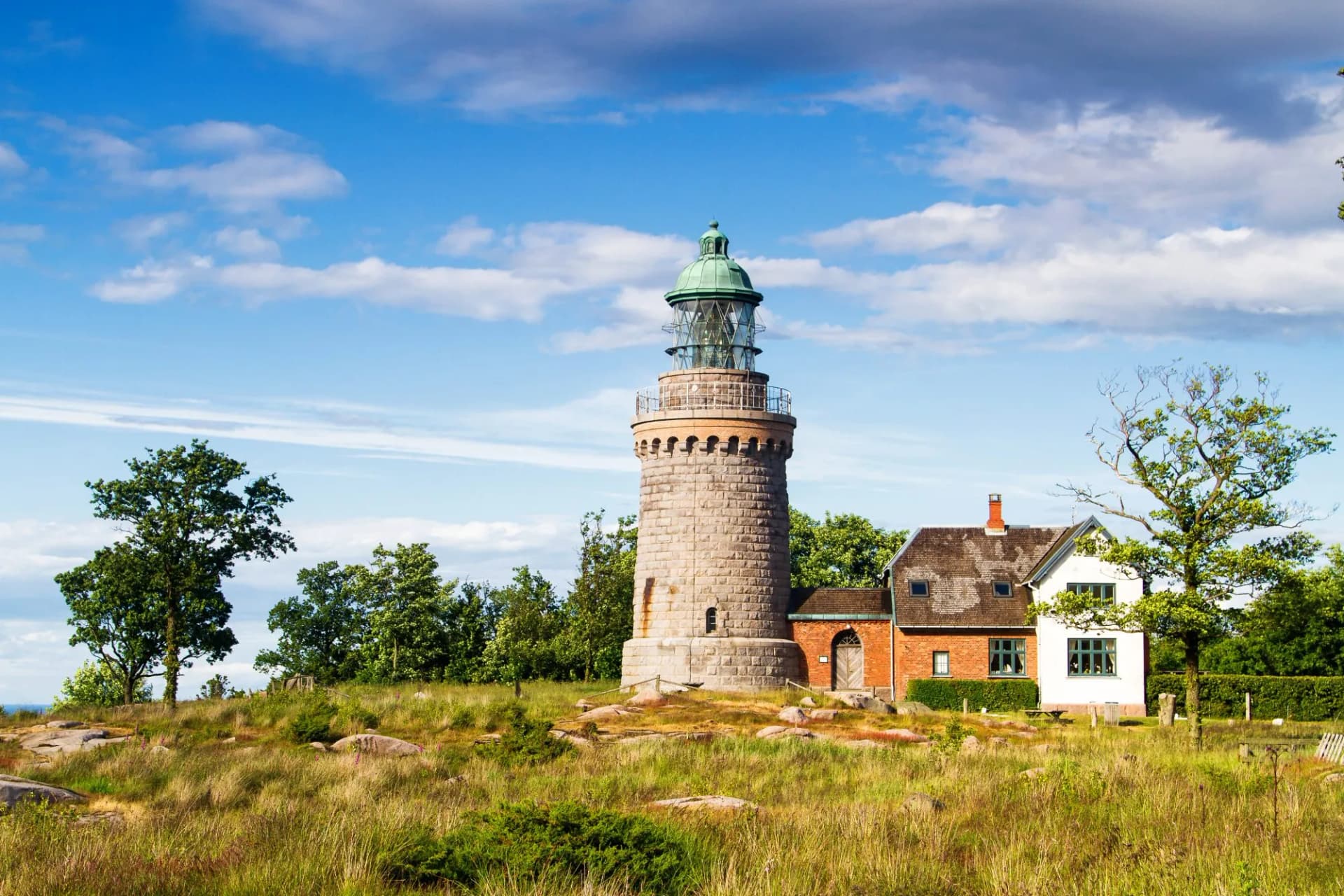 Hammeren Fyr lighthouse with stone tower and adjacent keeper's house on grassy hill under blue sky.