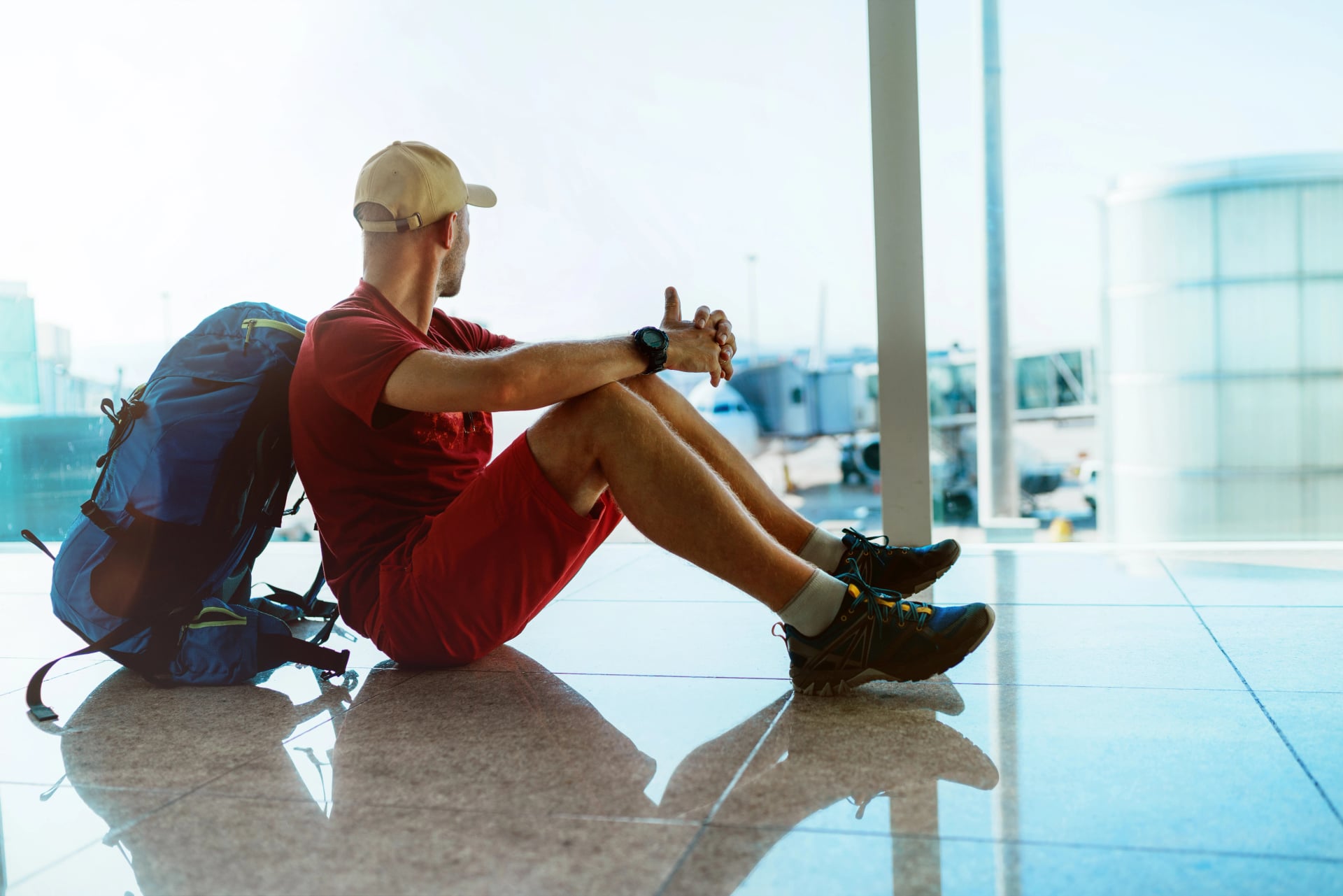 Alone backpacker traveller sitting on the airport terminal floor, looking at hall window and waiting for boarding at aircraft which prepared for flight behind a window