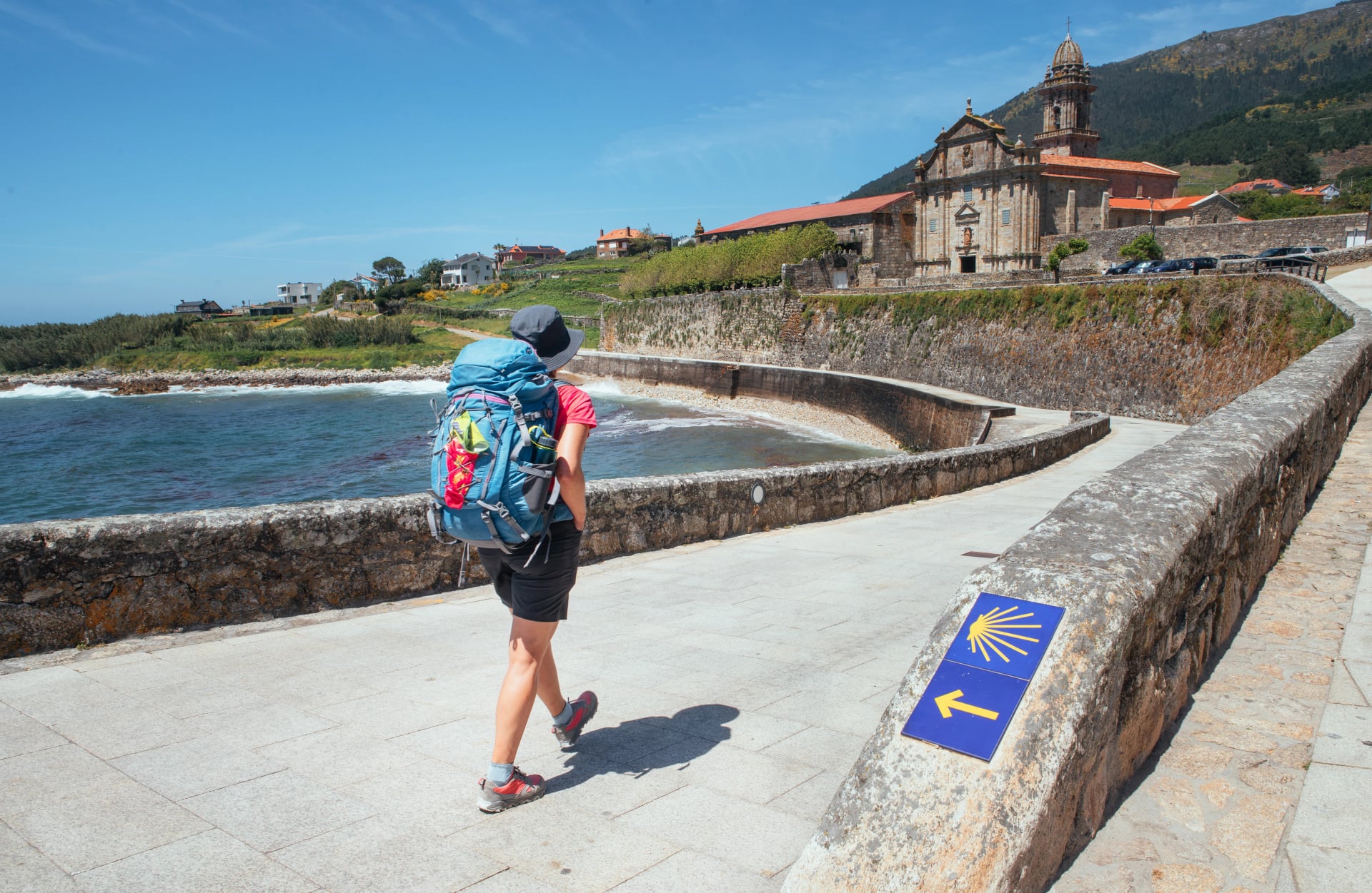 Female pilgrim with backpack walking next yellow arrow and shell directing Camino WAY inspiring solo walk on famous Camino de Santiago, Praia de Oia embankment, Royal Monastery of Santa María de Oia