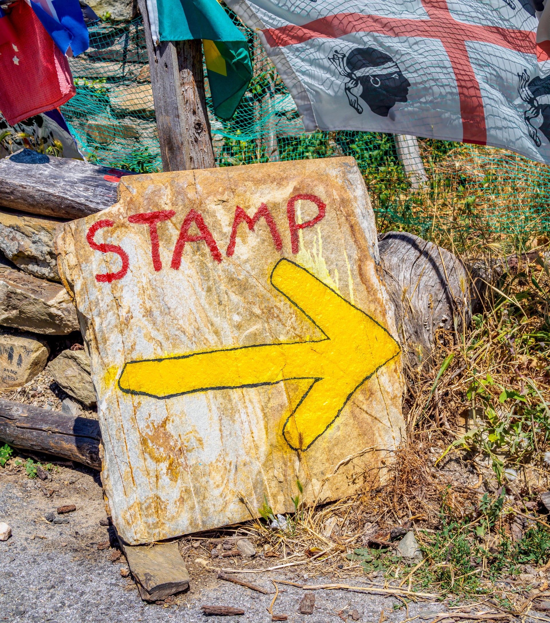 A weathered stone sign with a yellow arrow and the word STAMP, indicating a location to stamp the Compostela document on the Camino de Santiago, surrounded by mementos left by passing pilgrims.