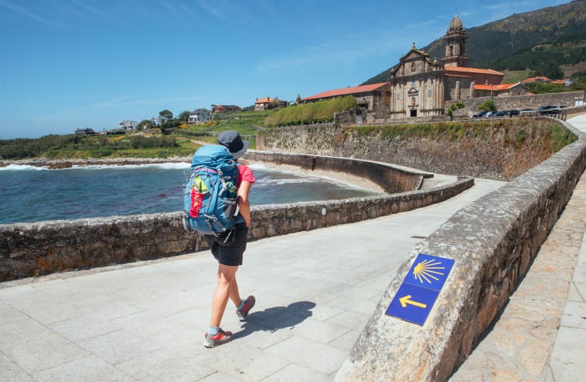 Female pilgrim with backpack walking next yellow arrow and shell directing Camino WAY inspiring solo walk on famous Camino de Santiago, Praia de Oia embankment, Royal Monastery of Santa María de Oia