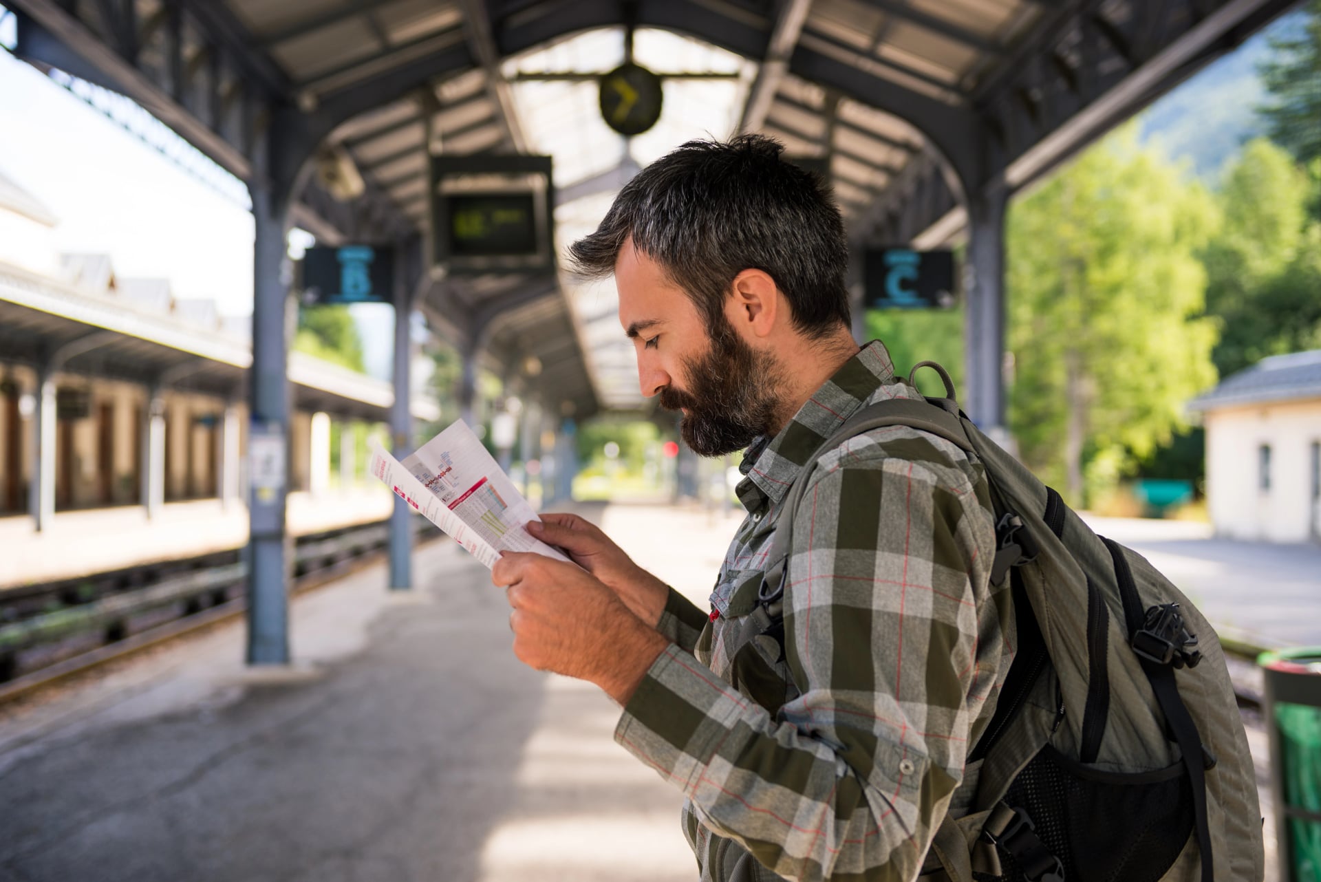 Young adult men commuter waiting for train in mountain train station reading timetable in sunny summer day.