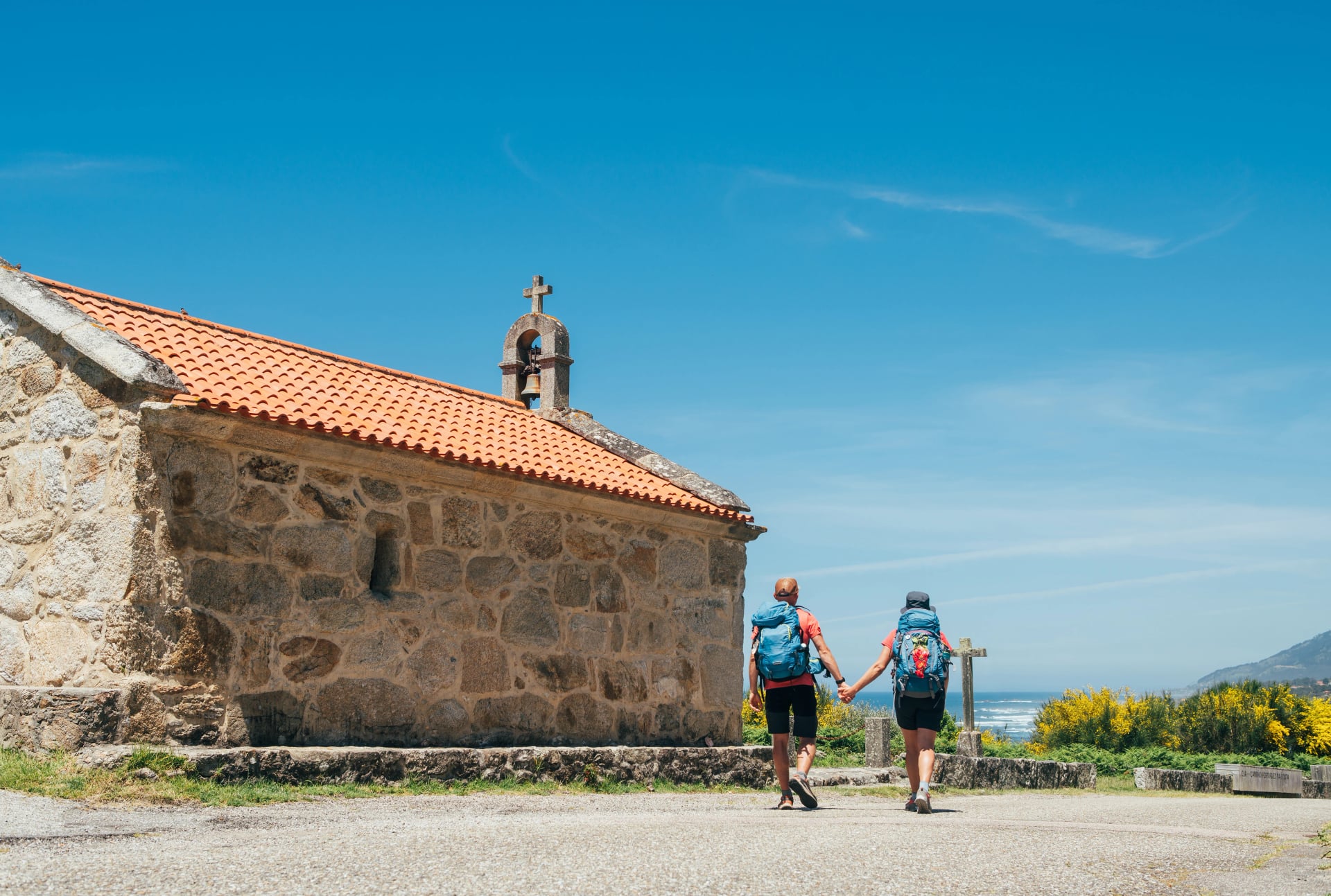 Couple of pilgrims with backpacks walking hand in hand next to old church on Camino Portuguese Way, inspiring walk on famous Camino de Santiago. Travel, pilgrimage, lifestyle, architecture