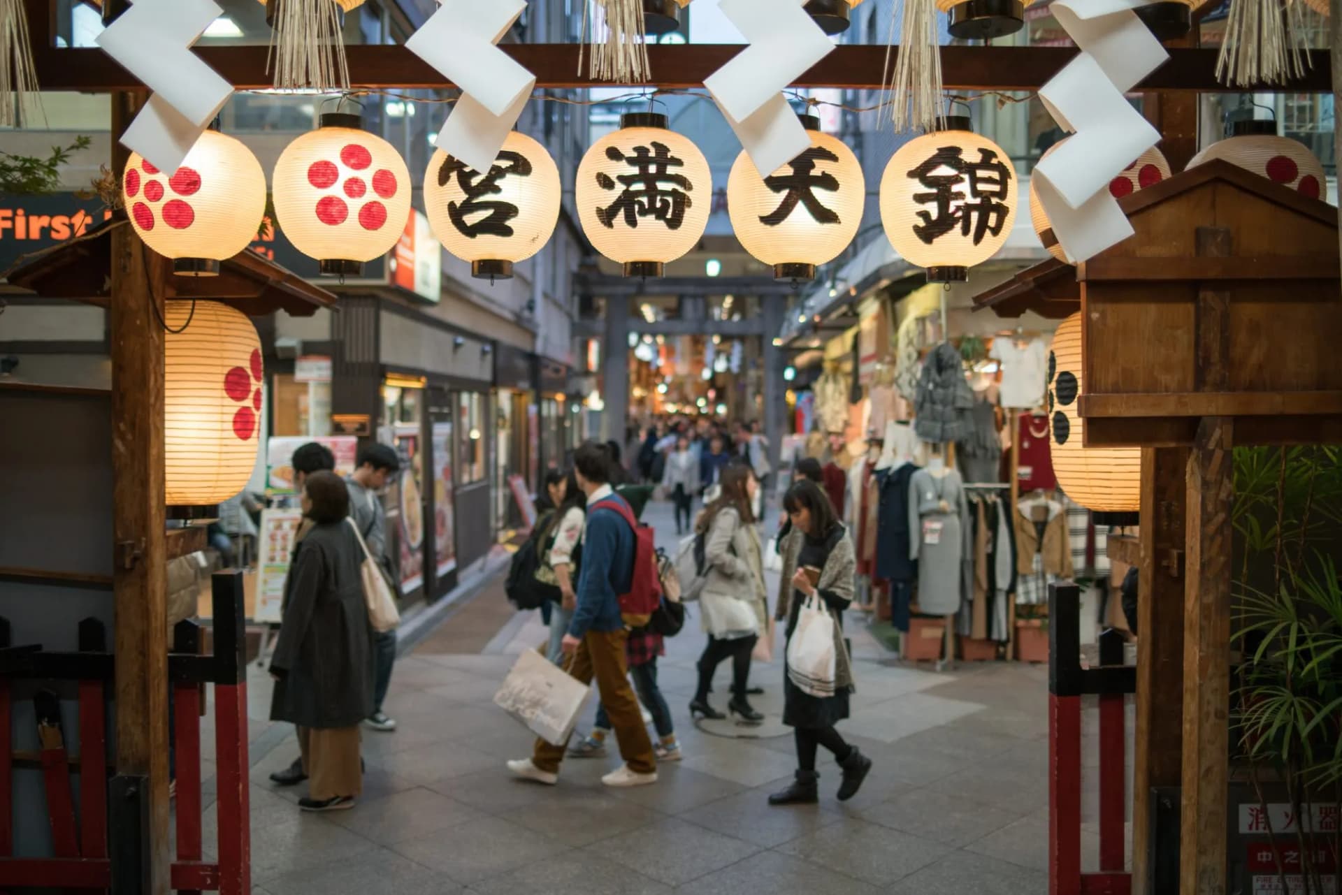 Lanterns hang over a shopping arcade with people walking toward a torii gate in Kyoto.