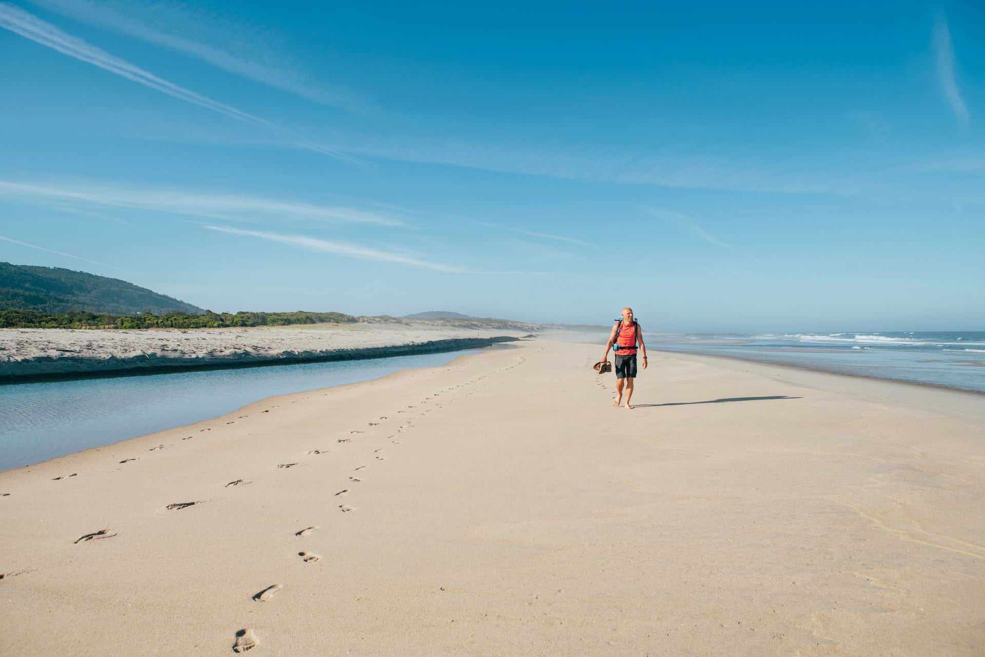 Lonely barefoot backpacker man walking by sandy Portugese beach in morning on the Atlantic ocean coast. Shot during the Camino Portuguese - popular pilgrimage route. Active people, traveling concept.