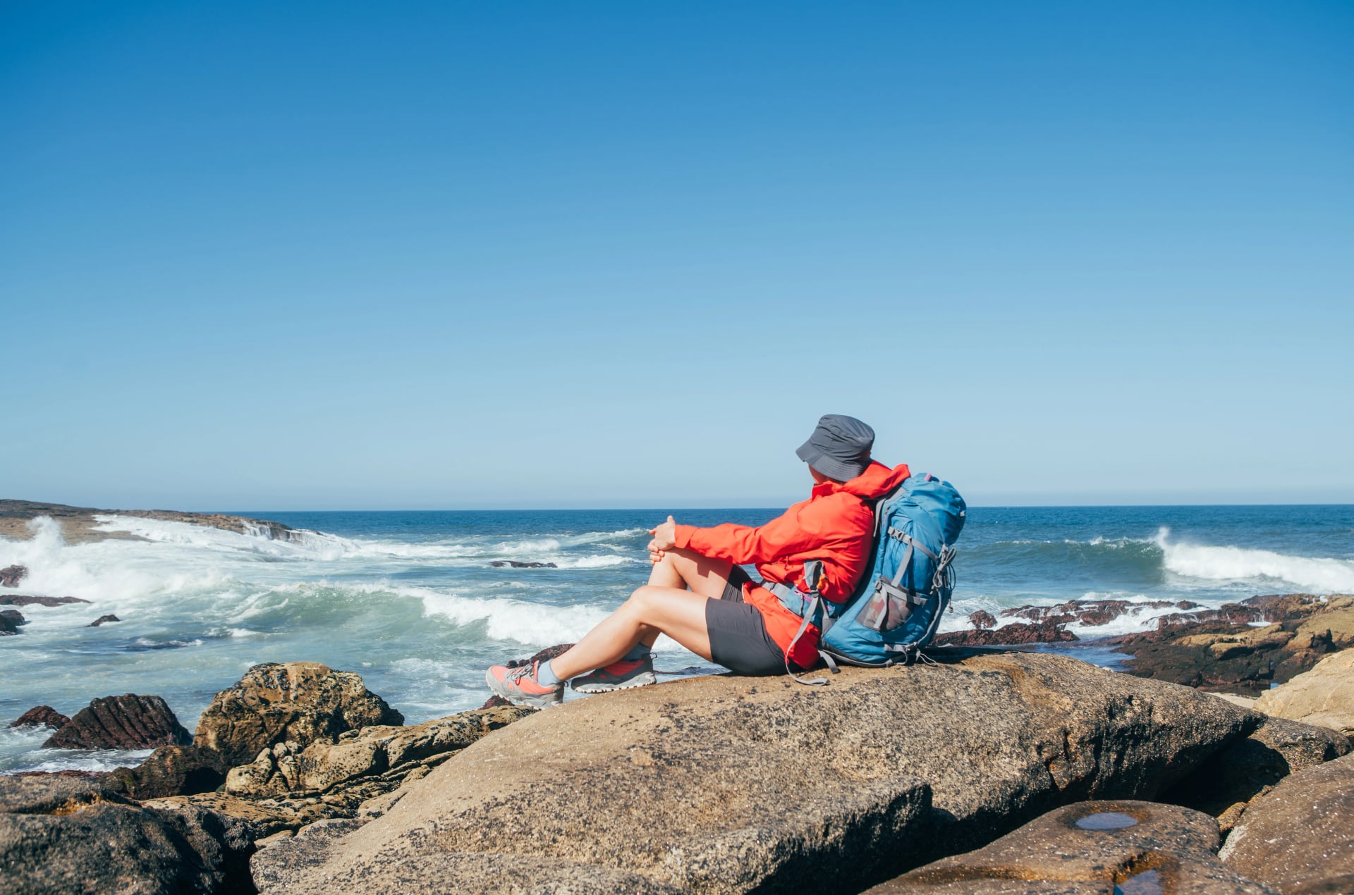 Beautiful short of female backpacker sitting on Atlantic ocean rocky coast in Spain enjoying a sea waves. Shot during the Camino Portuguese — popular pilgrimage route. Beauty in Nature concept.