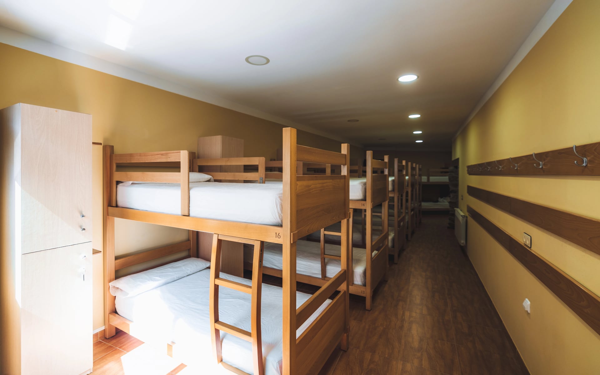 Clean and organized hostel dormitory room with multiple wooden bunk beds, individual lockers, wall hooks, and warm lighting, offering a functional shared sleeping space for travelers or pilgrims