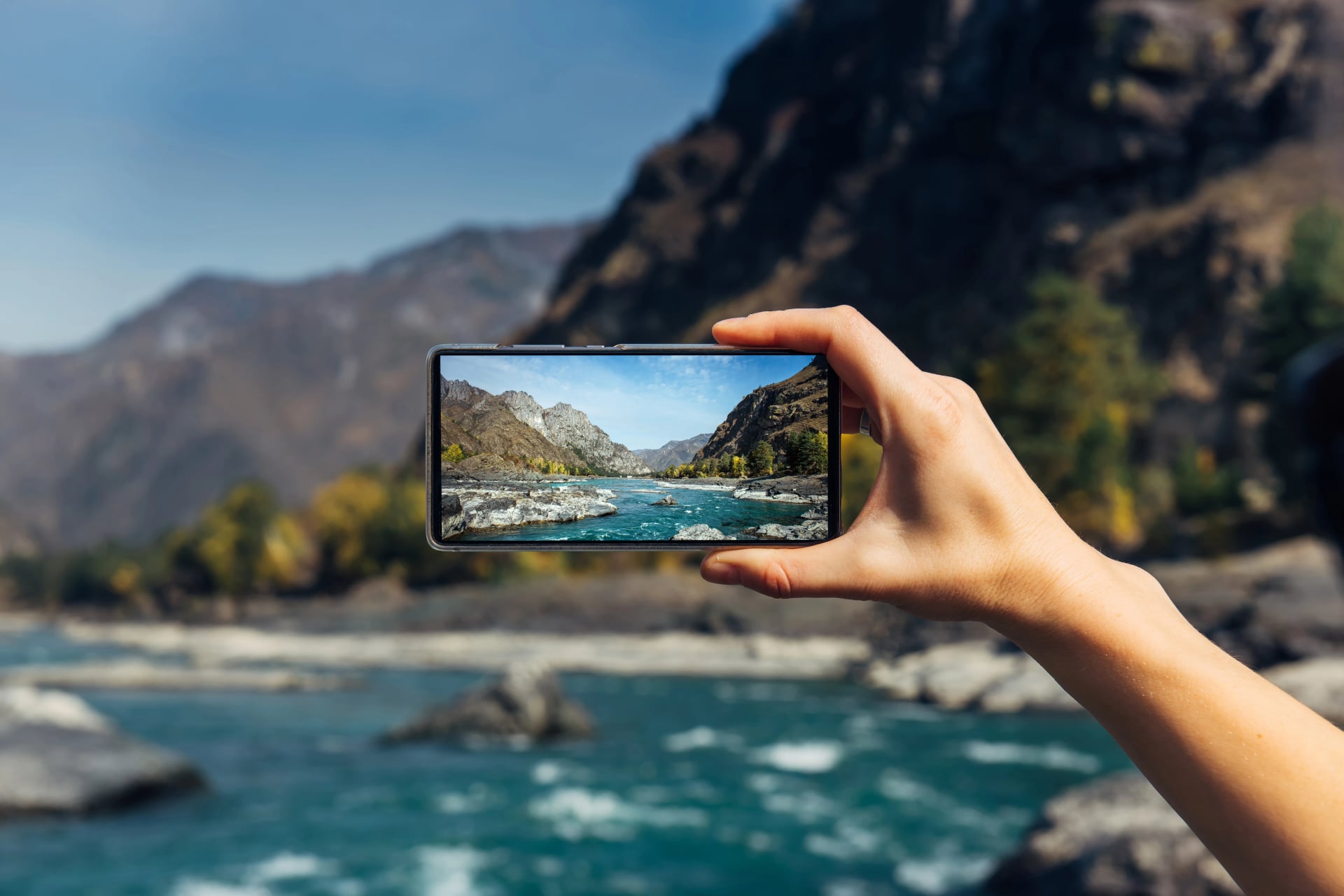 Female hand with smartphone close-up. Taking photo on smartphone while traveling. Delightful mountain landscape, stormy river, rocks and blue sky.