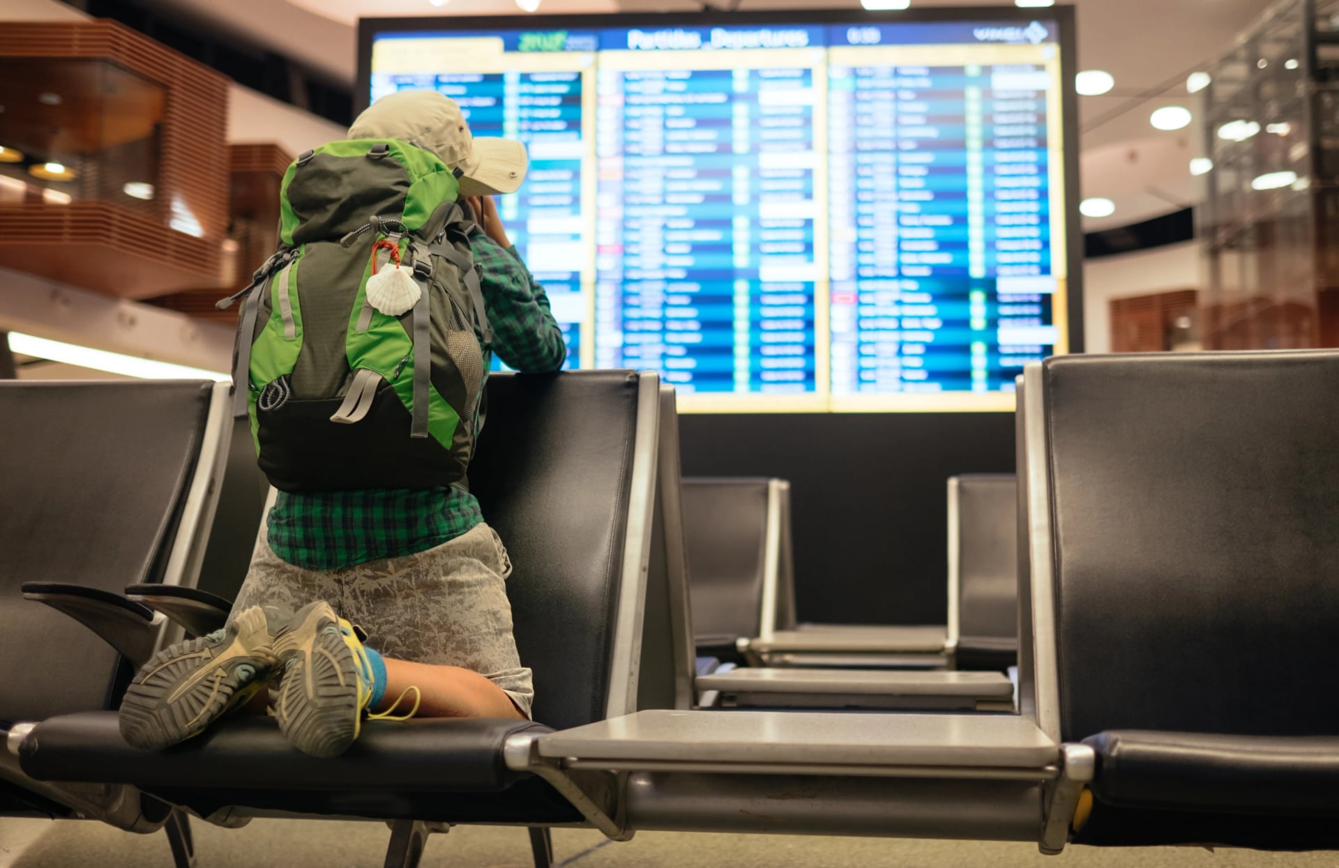 Young traveler boy with green backpack looking at airport departure board. Adventure vibe captures anticipation exploration, journey planning in modern terminal setting.Traveling and aerolines concept