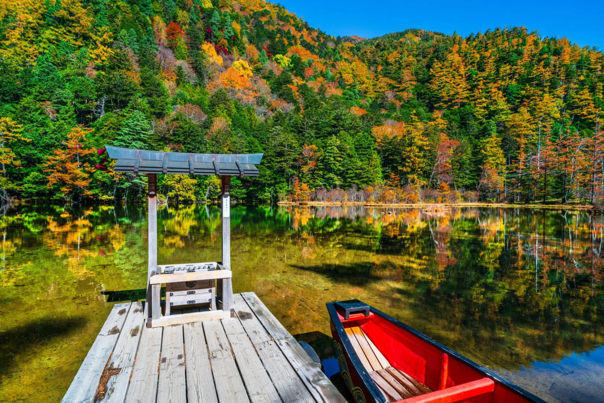 Wooden dock with offering box and red canoe on Myojin Pond with autumn foliage.