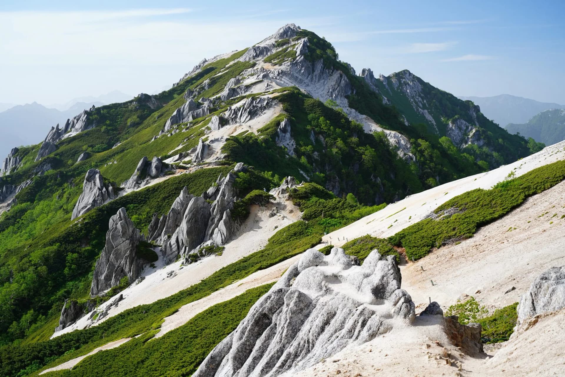 Rugged mountain ridge with green vegetation, light rock formations, and hiking trail under blue sky.
