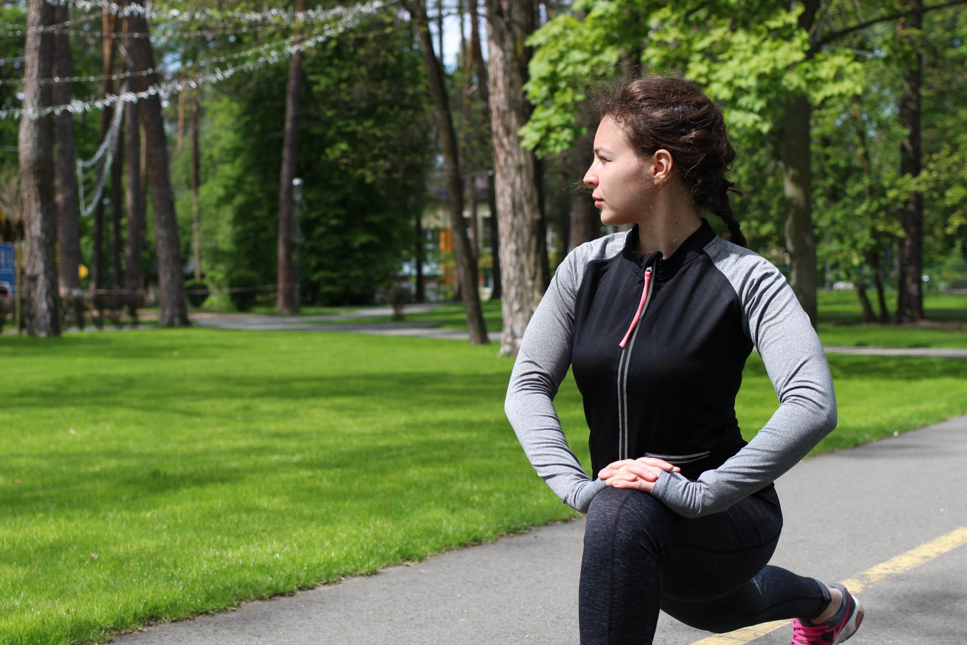 Young brunette athletic woman in grey sportswear stretching legs before running jogging in green park outdoor in sunny day.Concentrated millennial fitness girl doing lunges exercise,workout,training