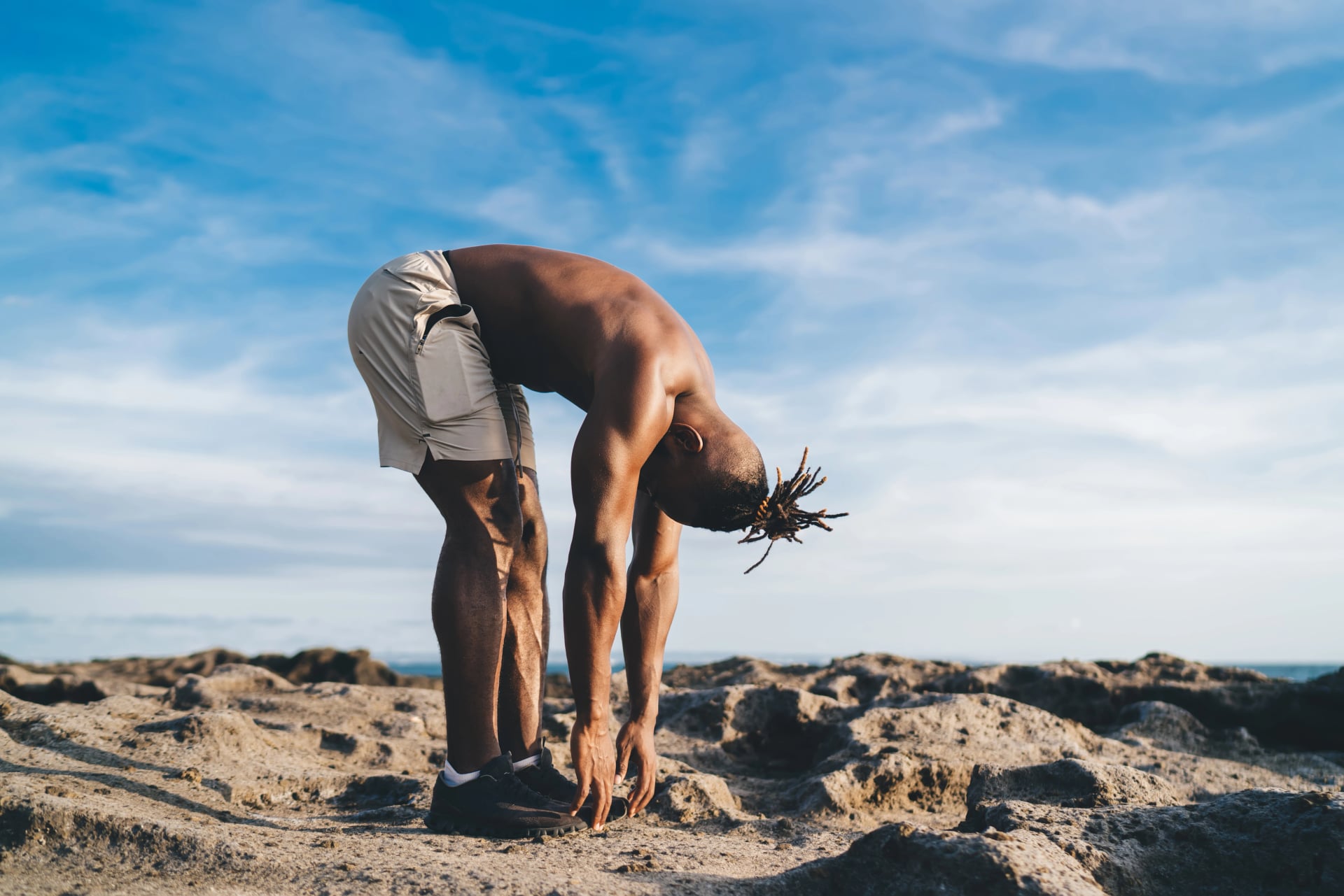 Unrecognizable black man doing standing forward bend exercise on rocky ground
