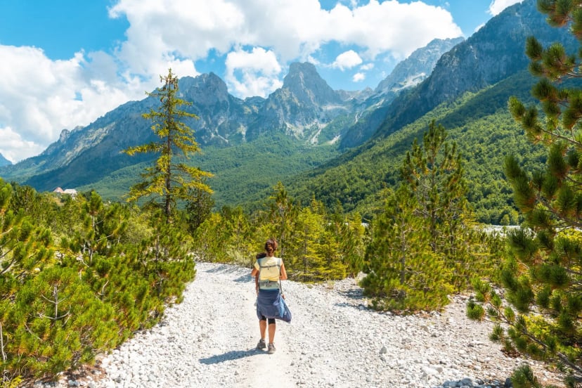 Woman hiking with child carrier on rocky path toward Valbona Valley mountains.