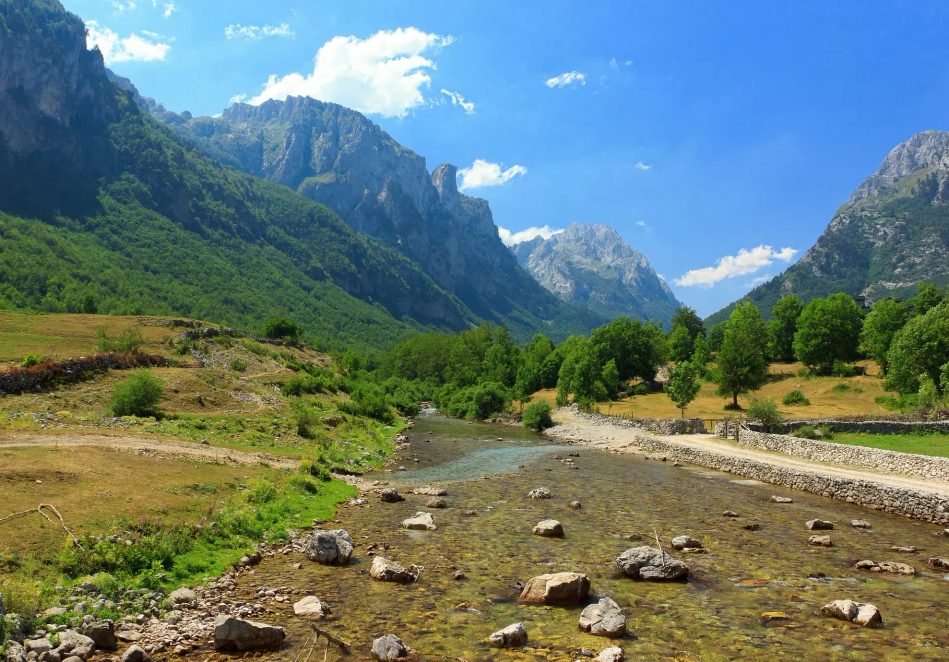 Clear river with rocks flowing through Ropojana valley surrounded by green mountains under blue sky.