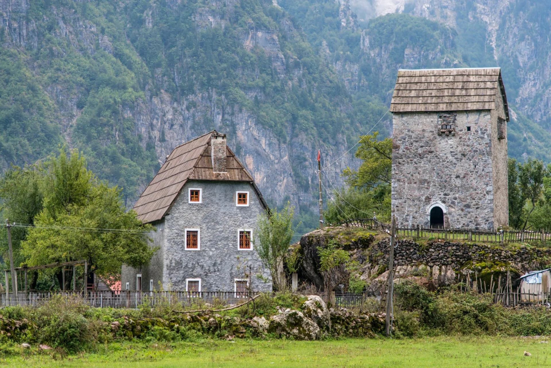 Koceku tower and stone house in Theth National Park against steep, forested mountains.