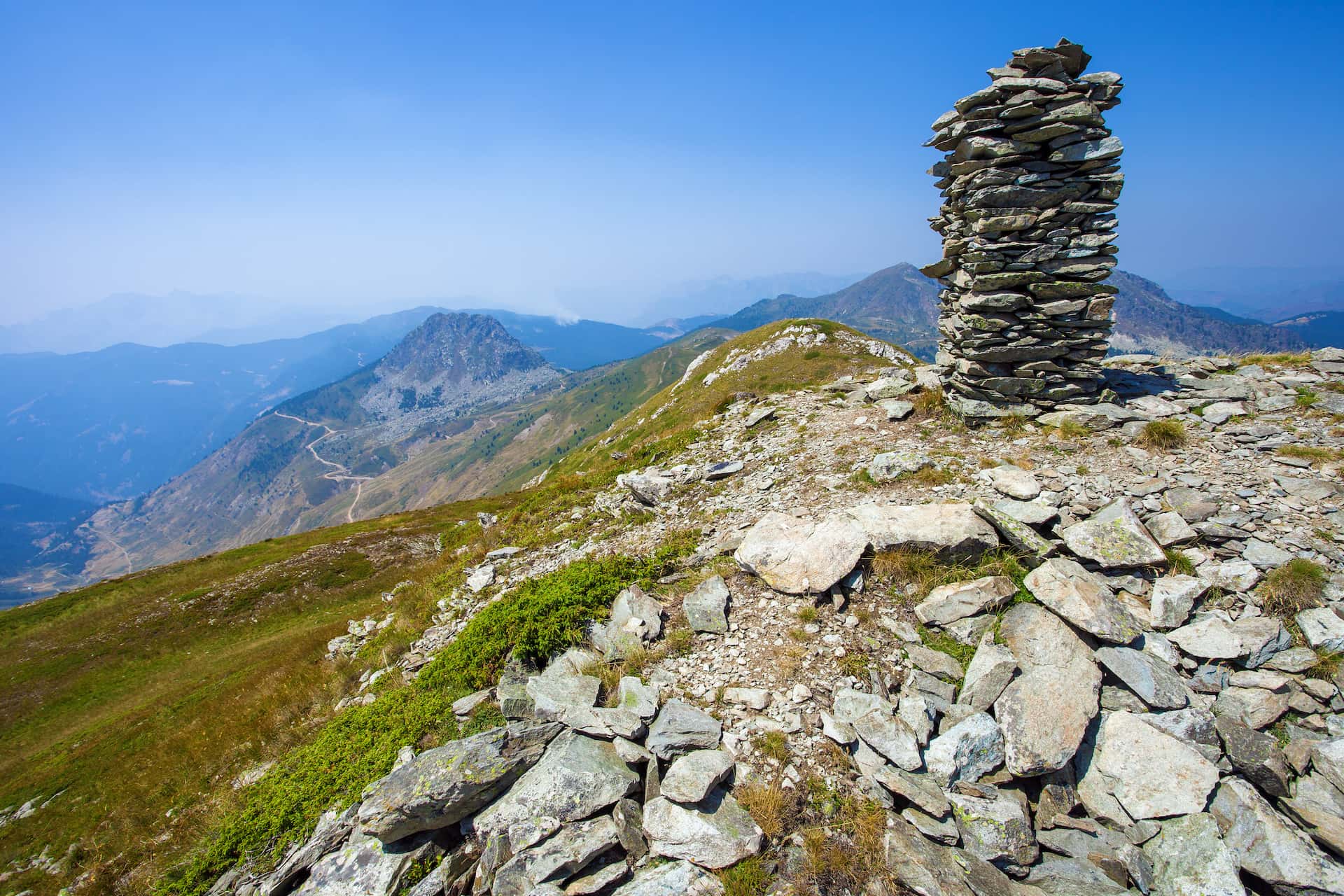 Stone cairn on rocky mountain ridge overlooking hazy blue valleys and winding road.