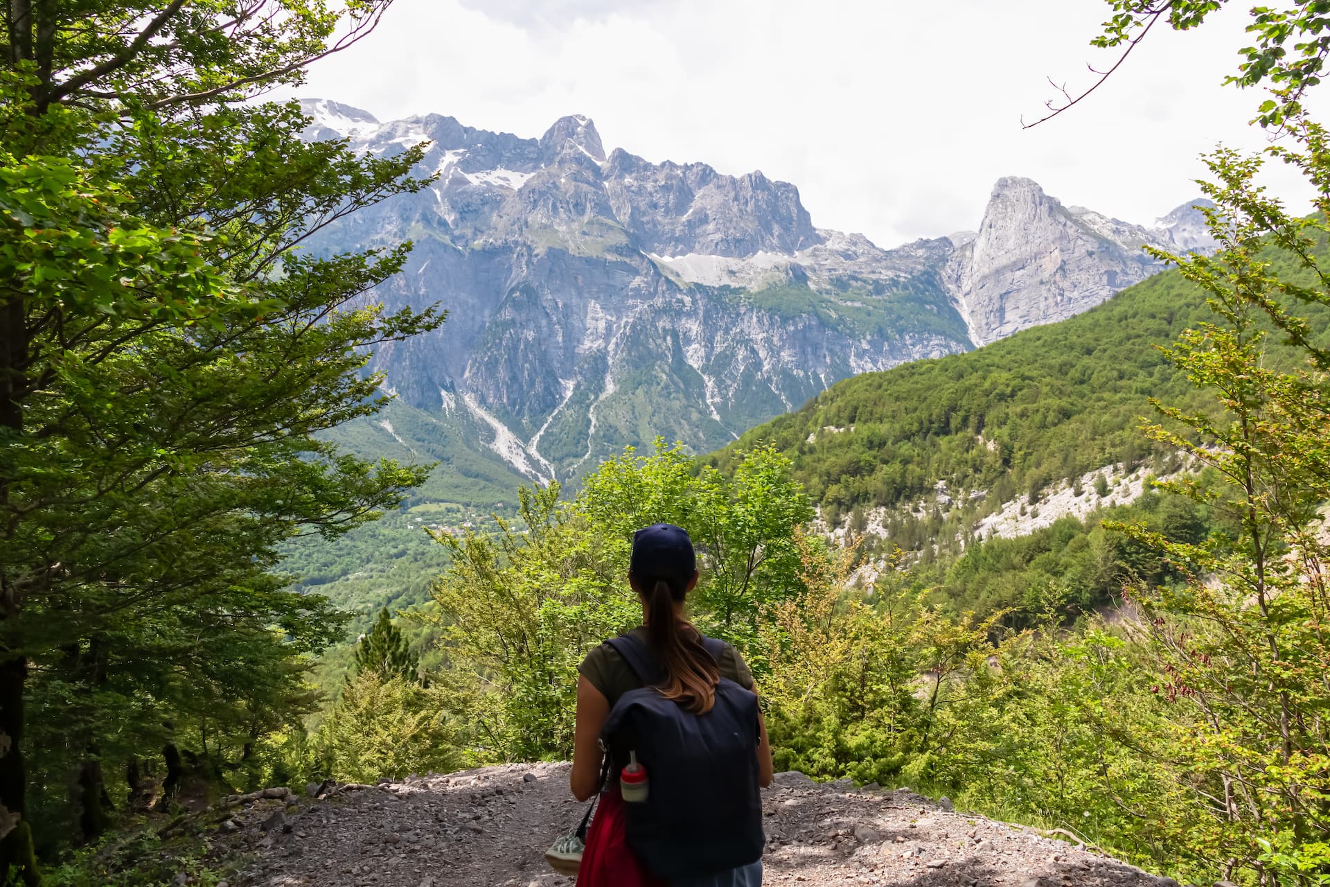 Hiker with backpack looking at snow-capped mountains and lush green valley in Albania.