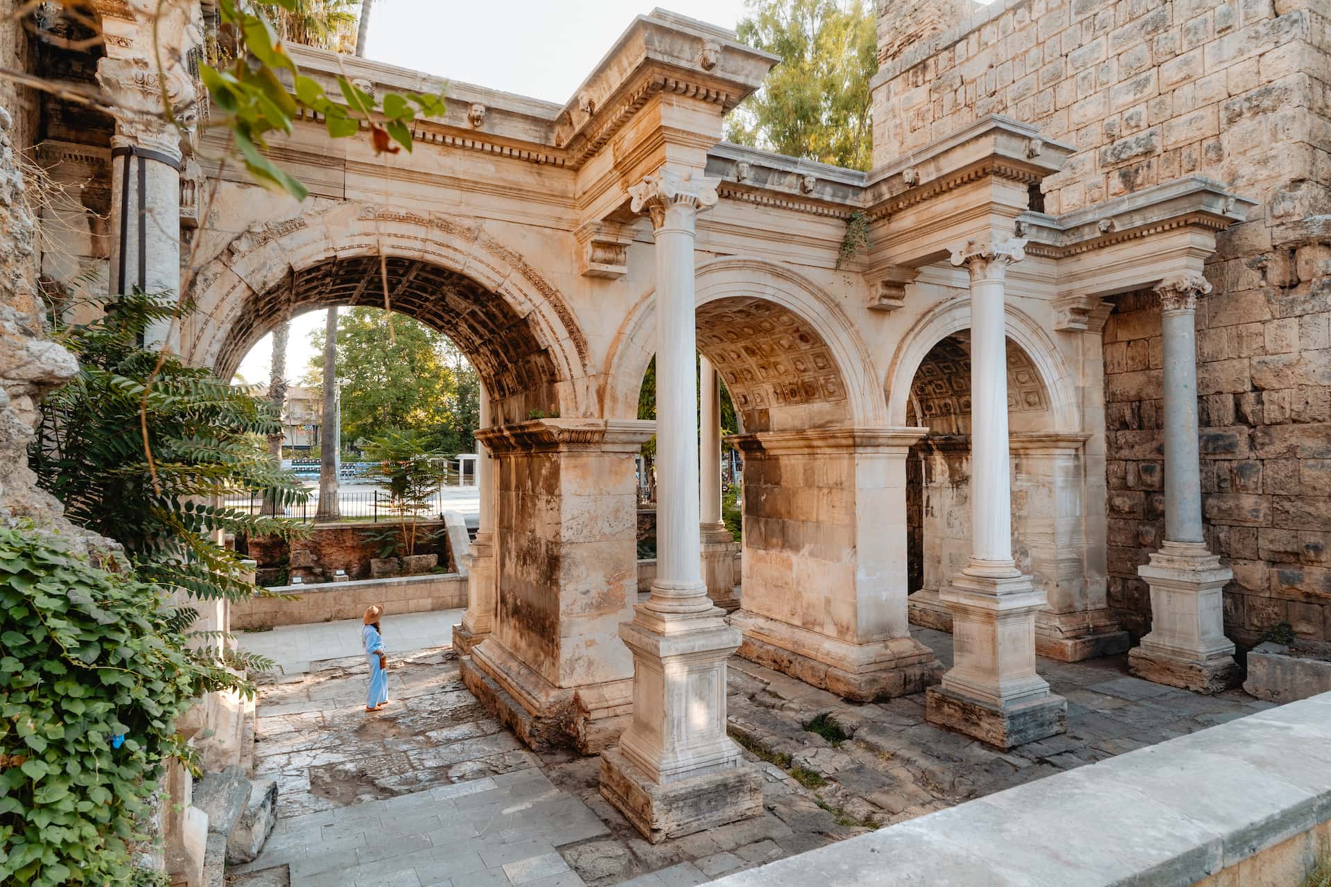 Ancient stone triumphal arch with columns, woman in sun hat standing below foliage.