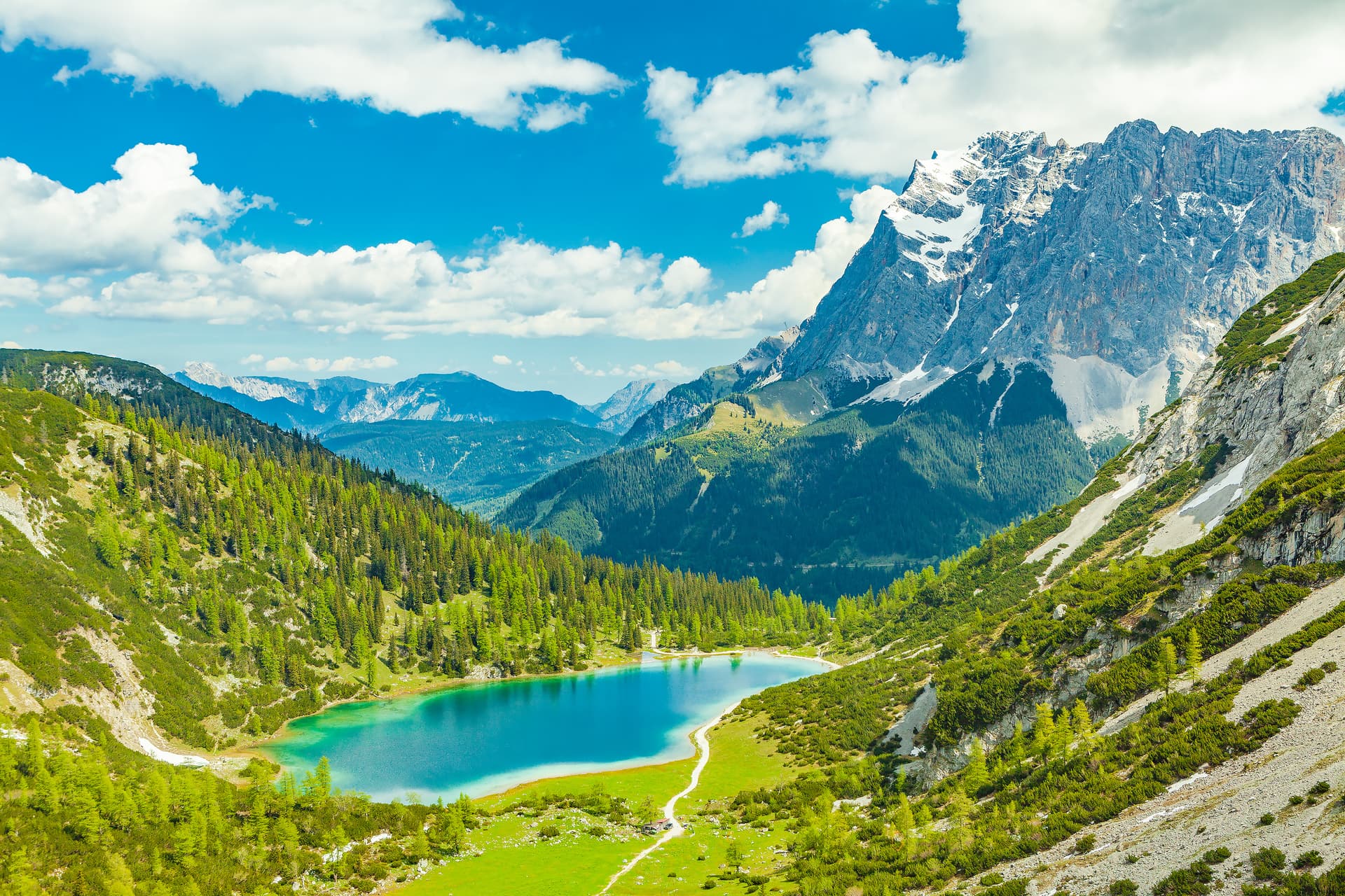Alpine lake with turquoise water surrounded by green slopes and snow-capped mountains under blue sky.
