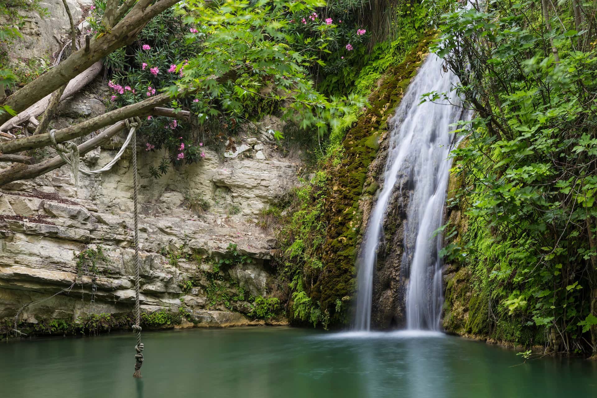 Waterfall cascading into a green pool with a rope swing by mossy cliffs in Cyprus.