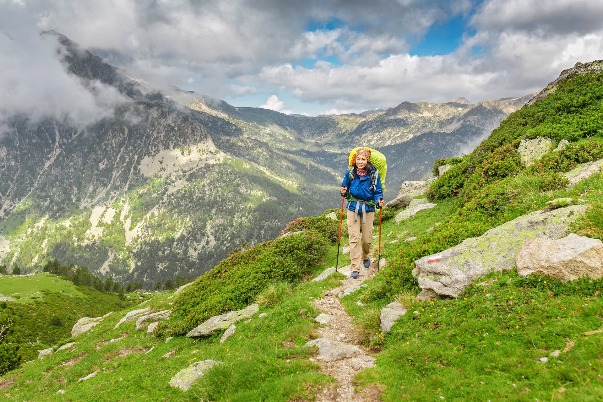 Hiker with backpack and poles trekking on grassy mountain trail with cloudy peaks in Andorra.