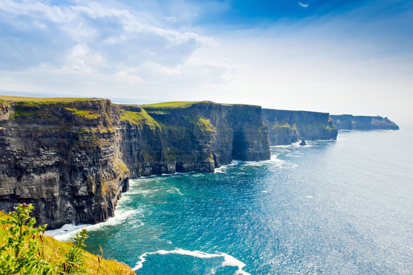 Towering Cliffs of Moher with green tops meeting bright blue ocean water under a partly cloudy sky.