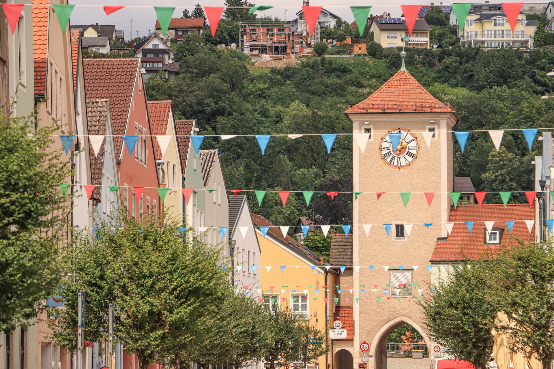 Clock tower in Kelheim center street decorated with colorful pennant banners and green trees.