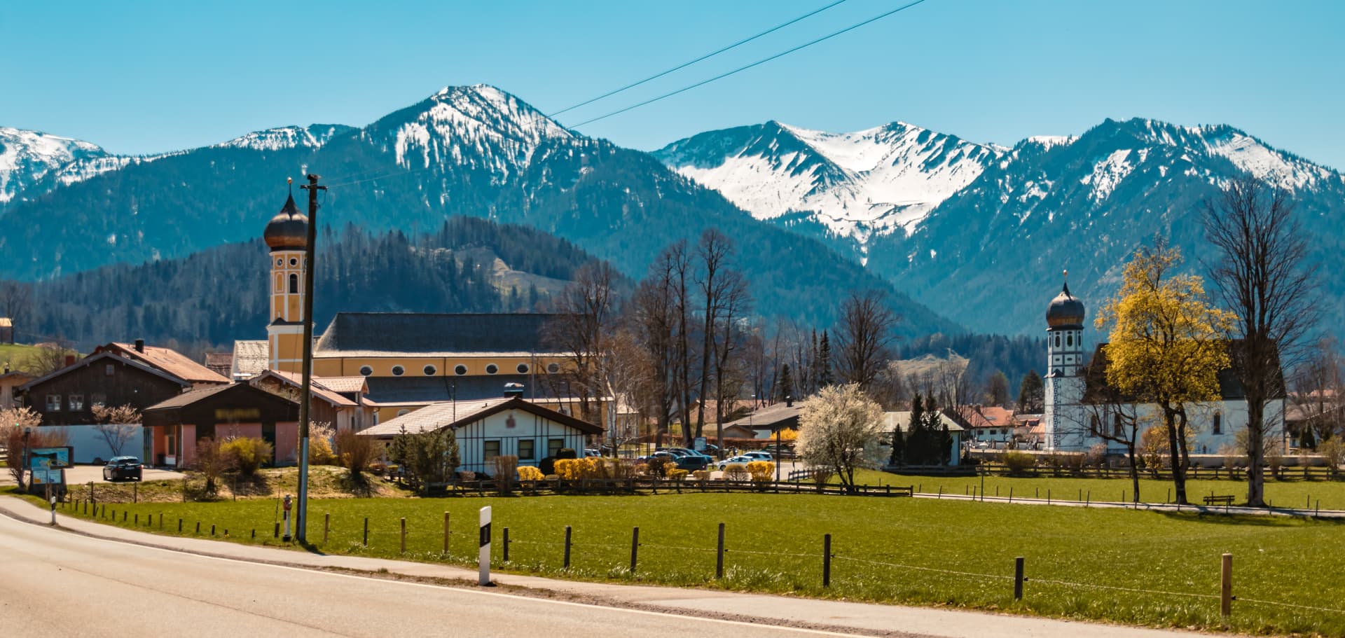 Alpine village with church towers set against snow-capped mountains under a clear blue sky.