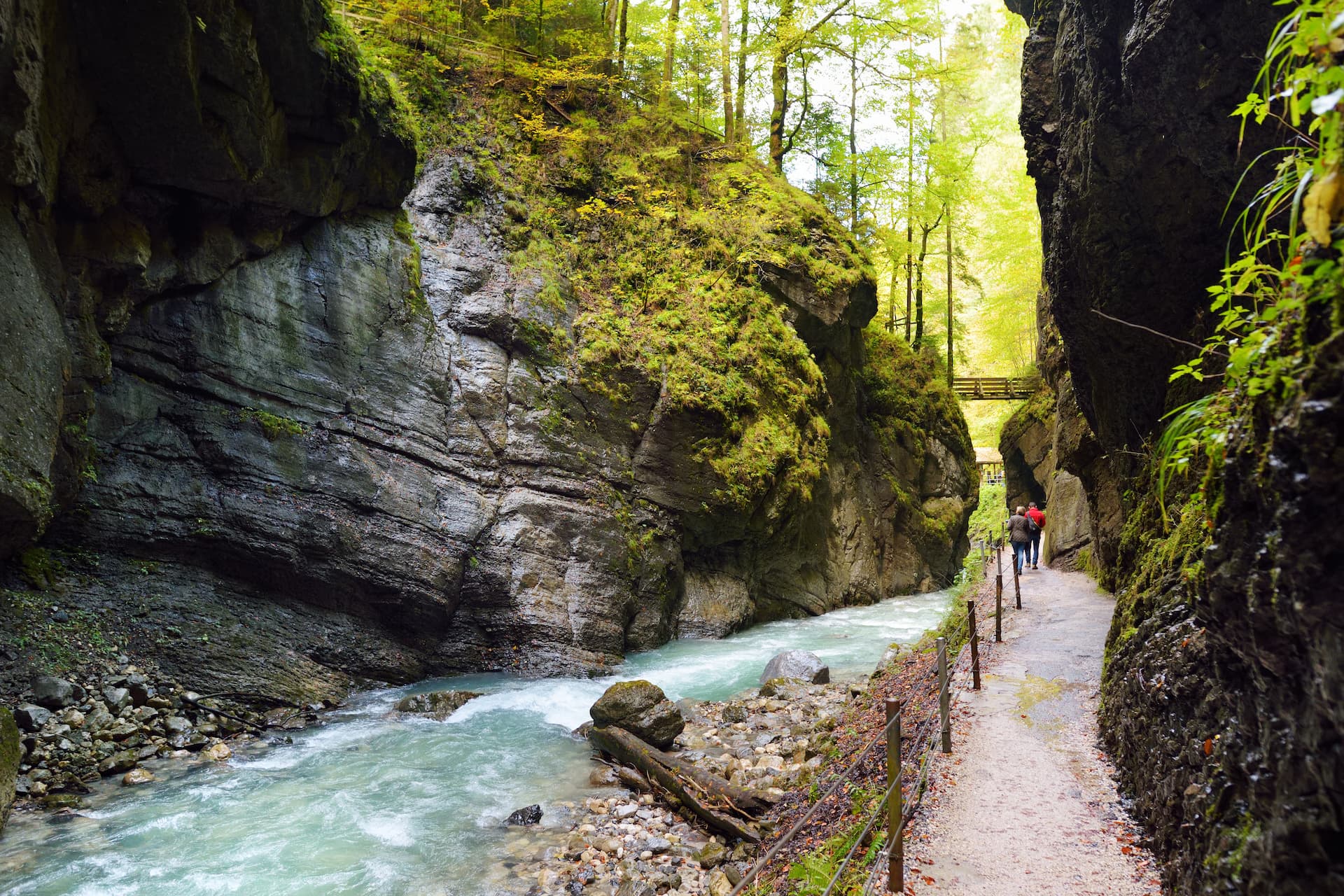Hikers on path beside turquoise river in mossy Partnach Gorge with wooden bridge overhead.