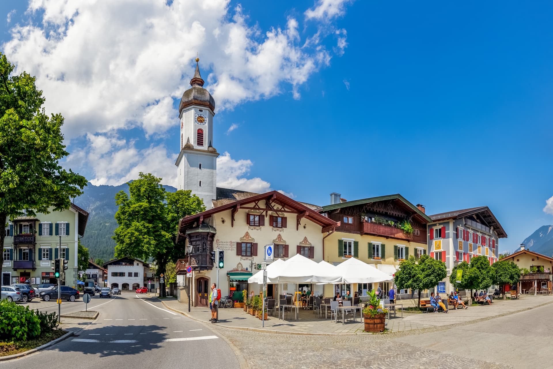 Town square in Garmisch-Partenkirchen with church tower, outdoor cafe, and alpine mountains.